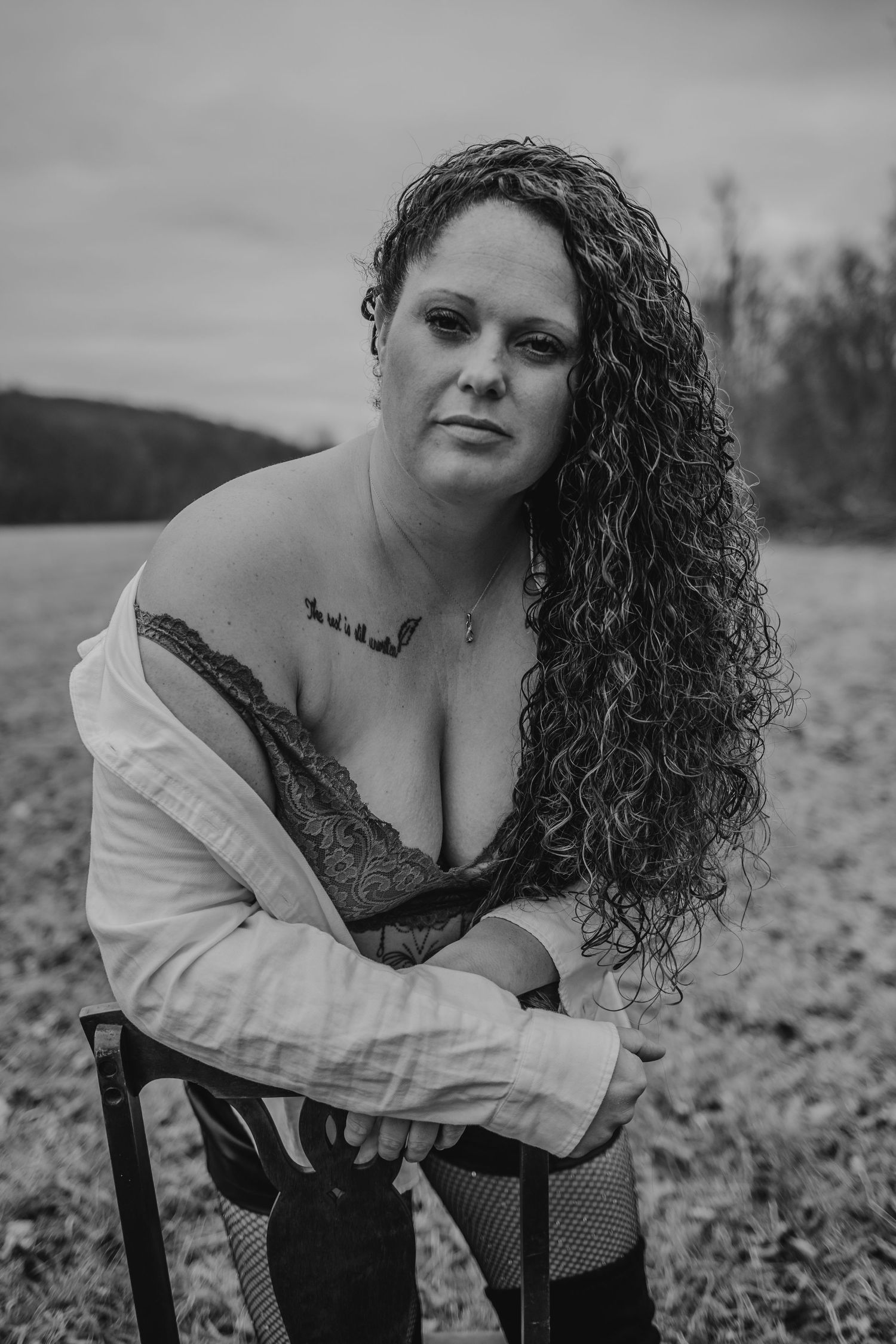 Black and white portrait of a curly-haired woman in an open shirt and lace bra sitting on a chair outdoors.