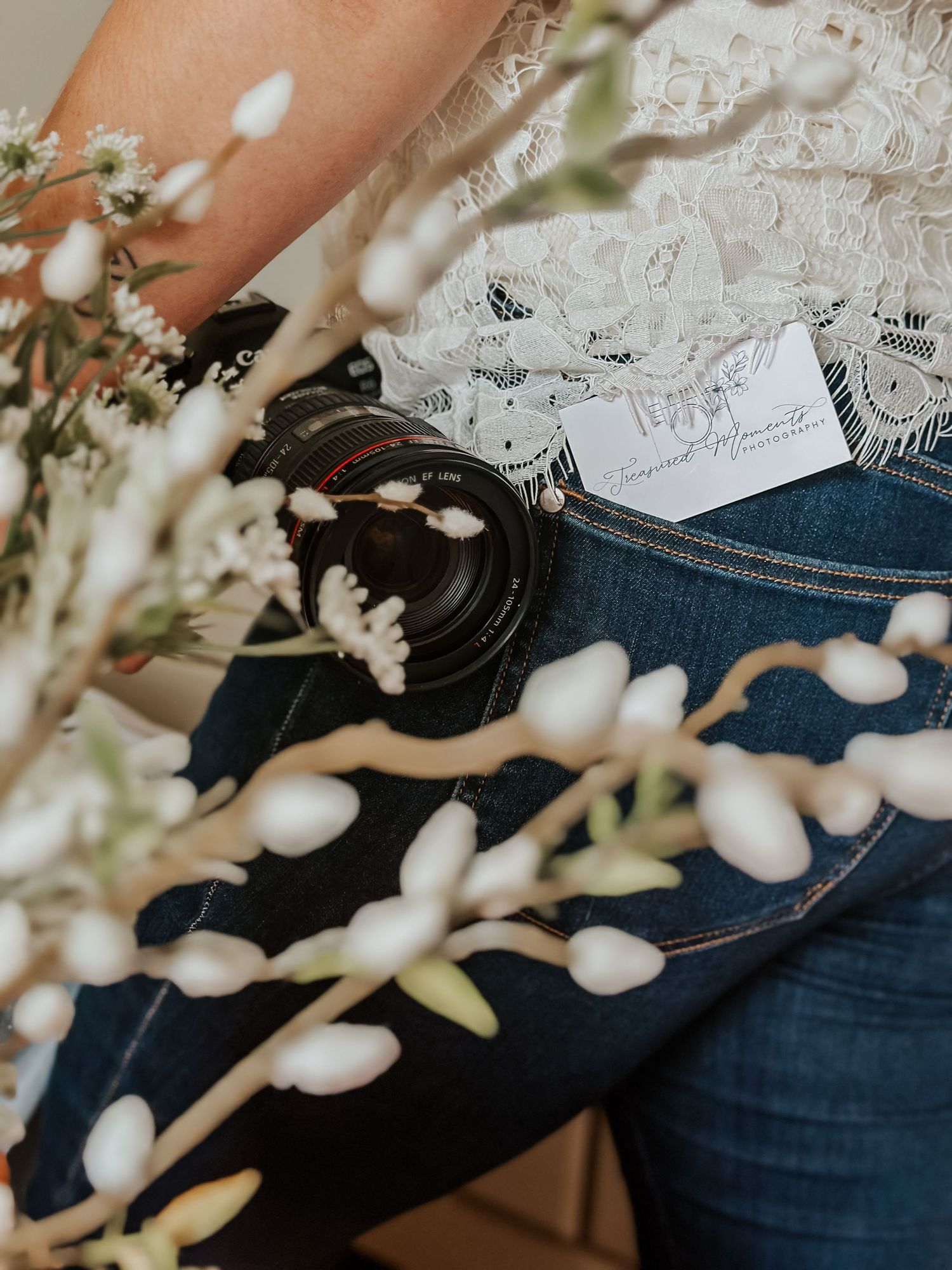 Person in lace top and jeans holding a Canon camera with white wildflowers in the foreground.