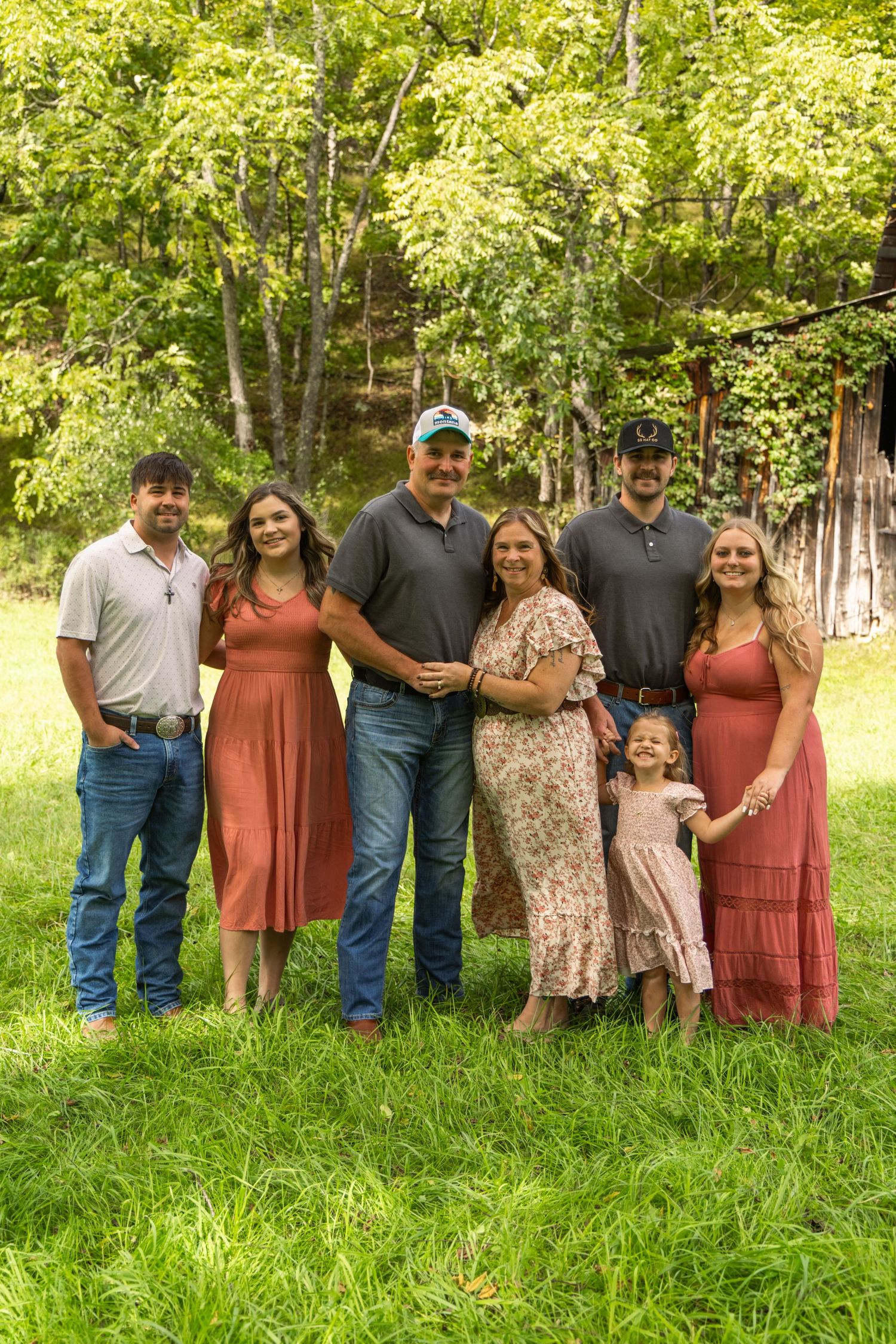 Family of seven in coordinated outfits, including a young girl, smiling together on a green lawn in front of trees and a rustic barn.