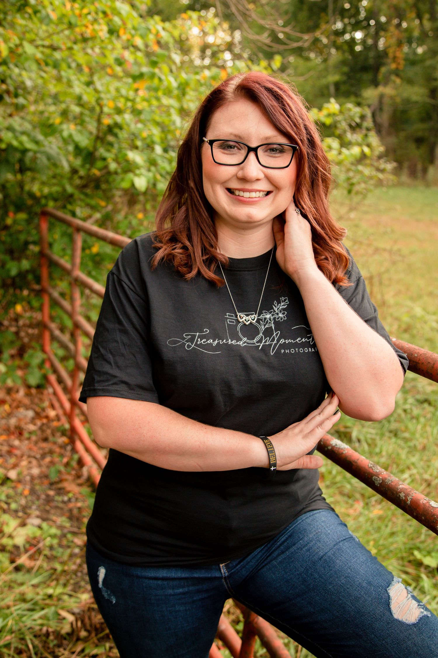 Smiling redhead woman with glasses wearing a black Treasured Moments Photography shirt, leaning on a rustic fence outdoors.