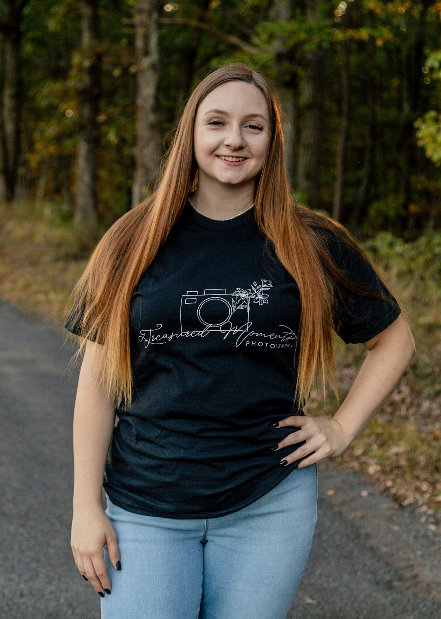 Young woman with long auburn hair smiling outdoors, wearing a black photography logo t-shirt and jeans on a tree-lined path.