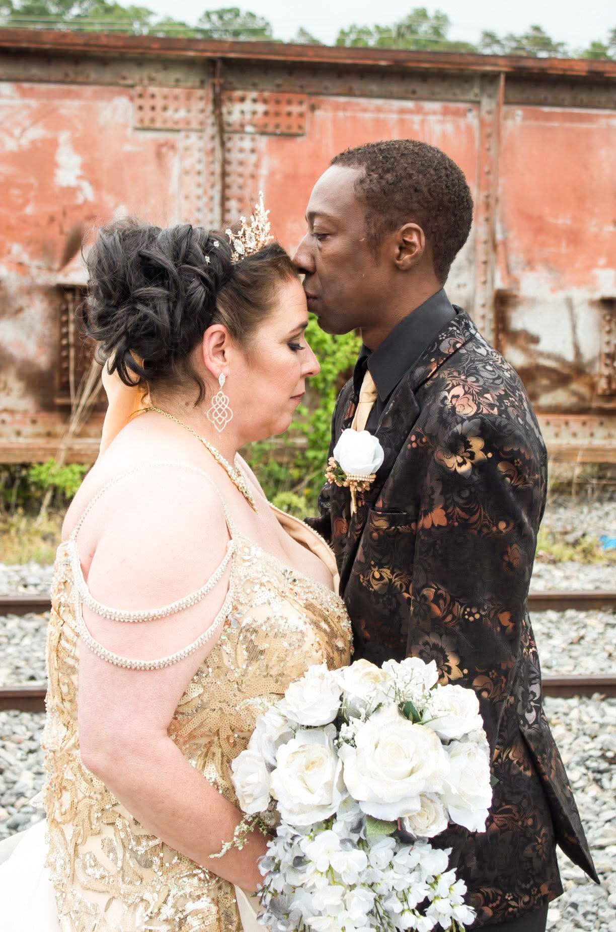 Interracial couple in wedding attire sharing a tender moment near rustic railroad tracks, bride holding white rose bouquet.
