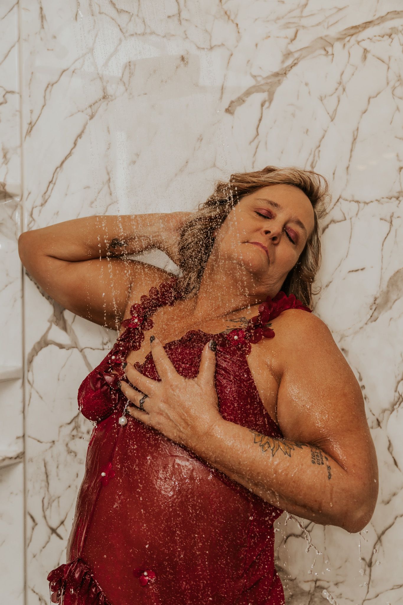 A woman in a red sequin dress poses against a marble wall while water cascades over her.