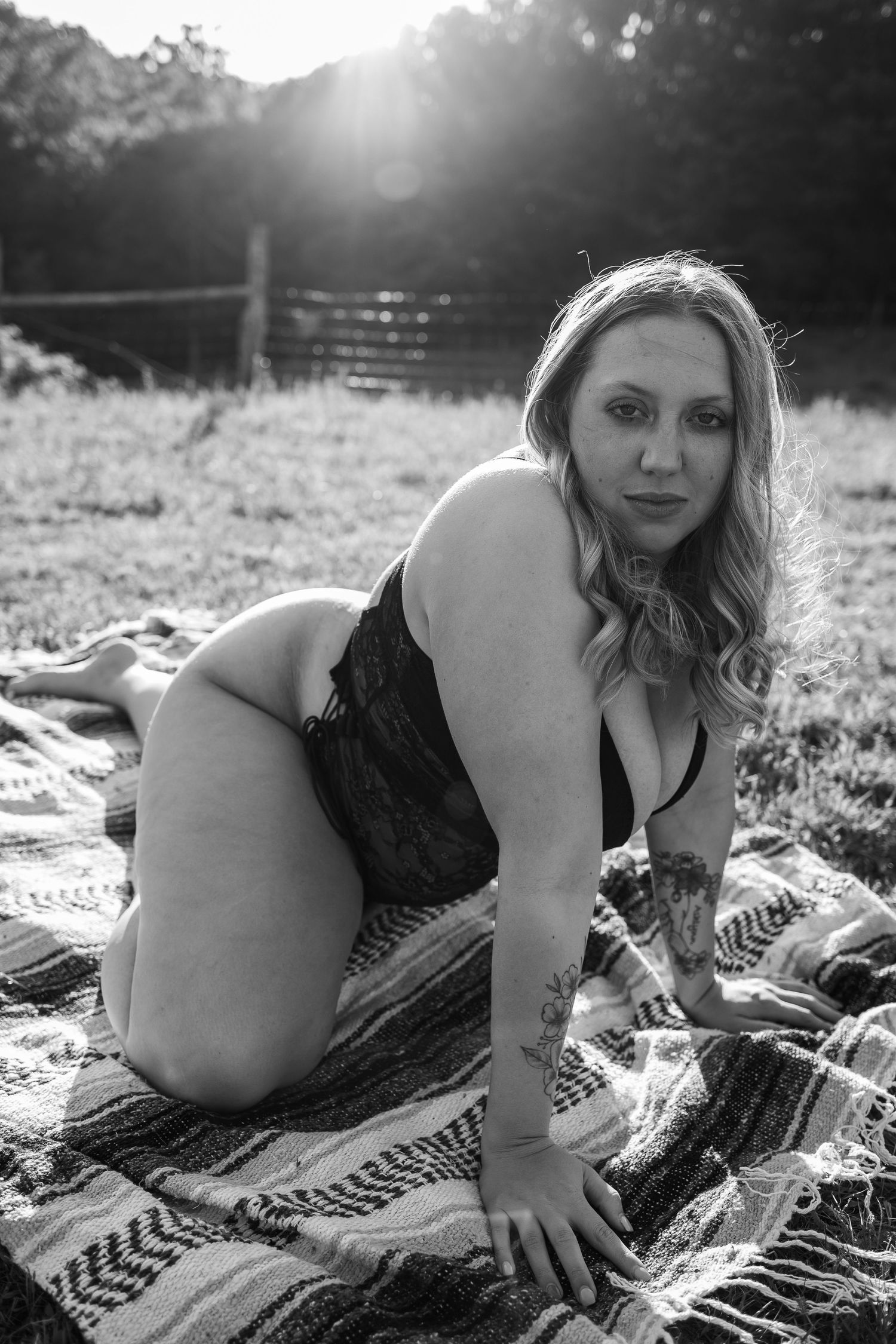 Black and white photo of a tattooed blonde woman in black lace lingerie posing on a blanket in a sunny field.
