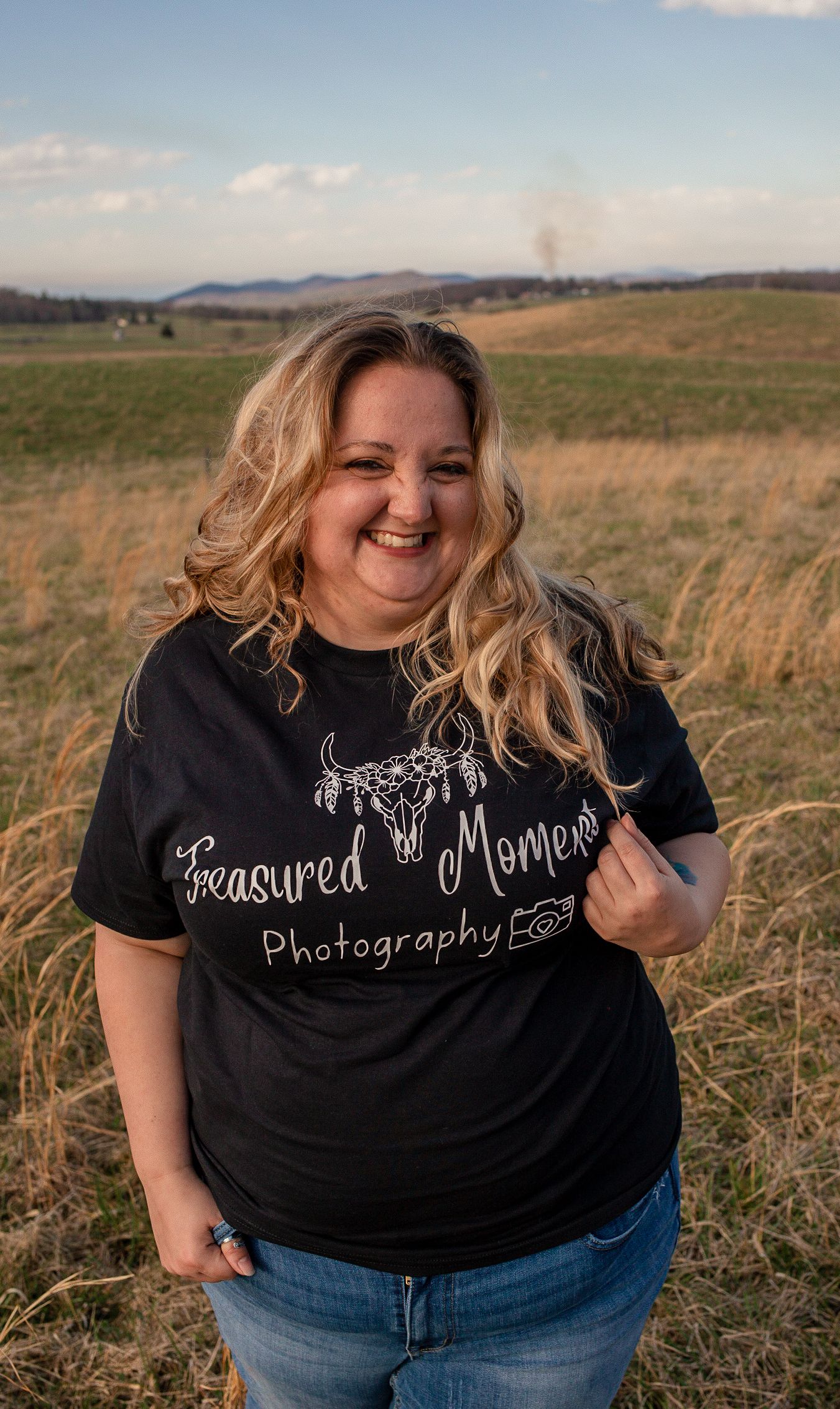 Smiling blonde woman wearing a black Treasured Moments Photography shirt standing in an open field at sunset.