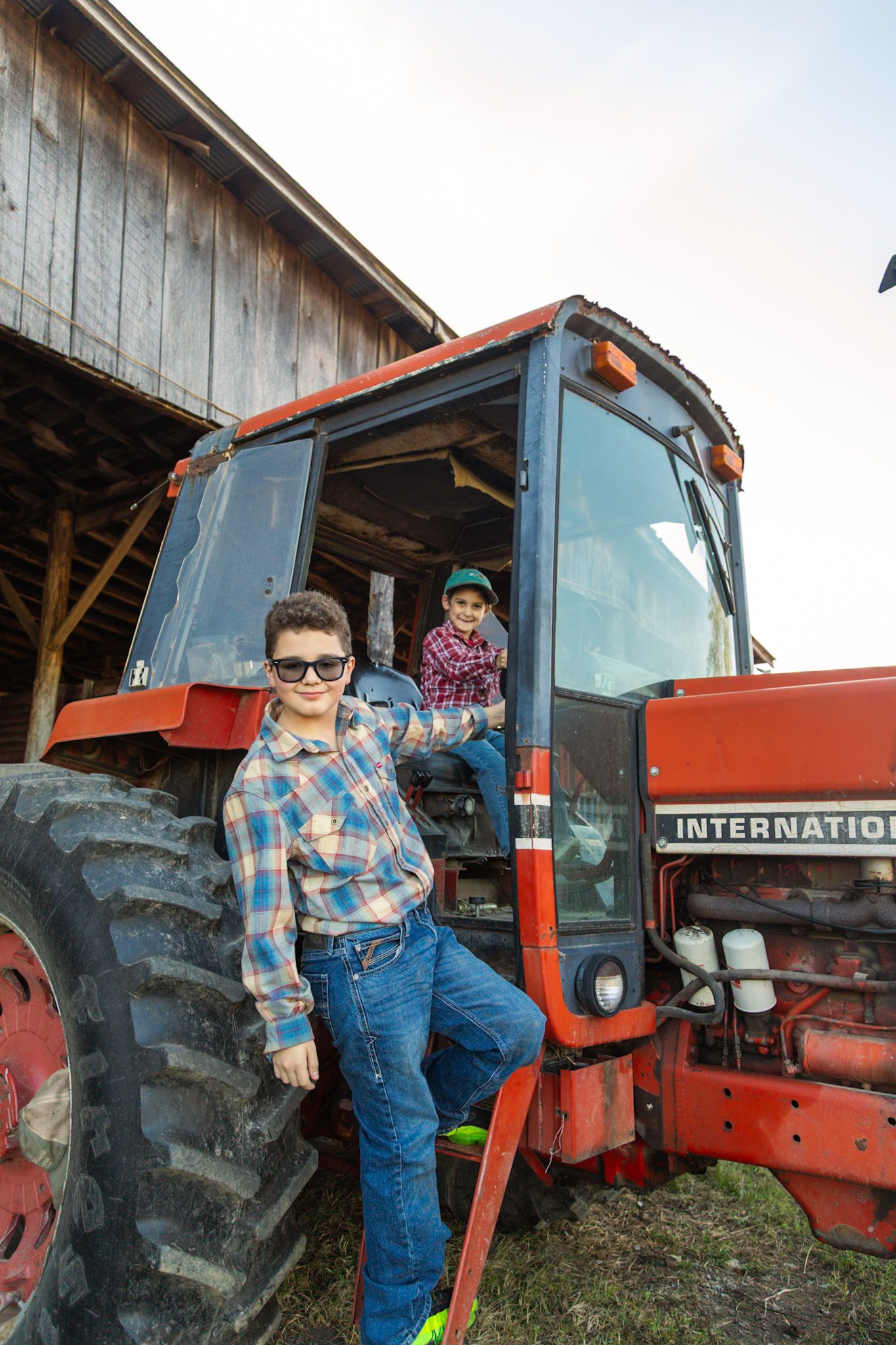 Two kids in plaid shirts pose with red International tractor near rustic wooden barn.
