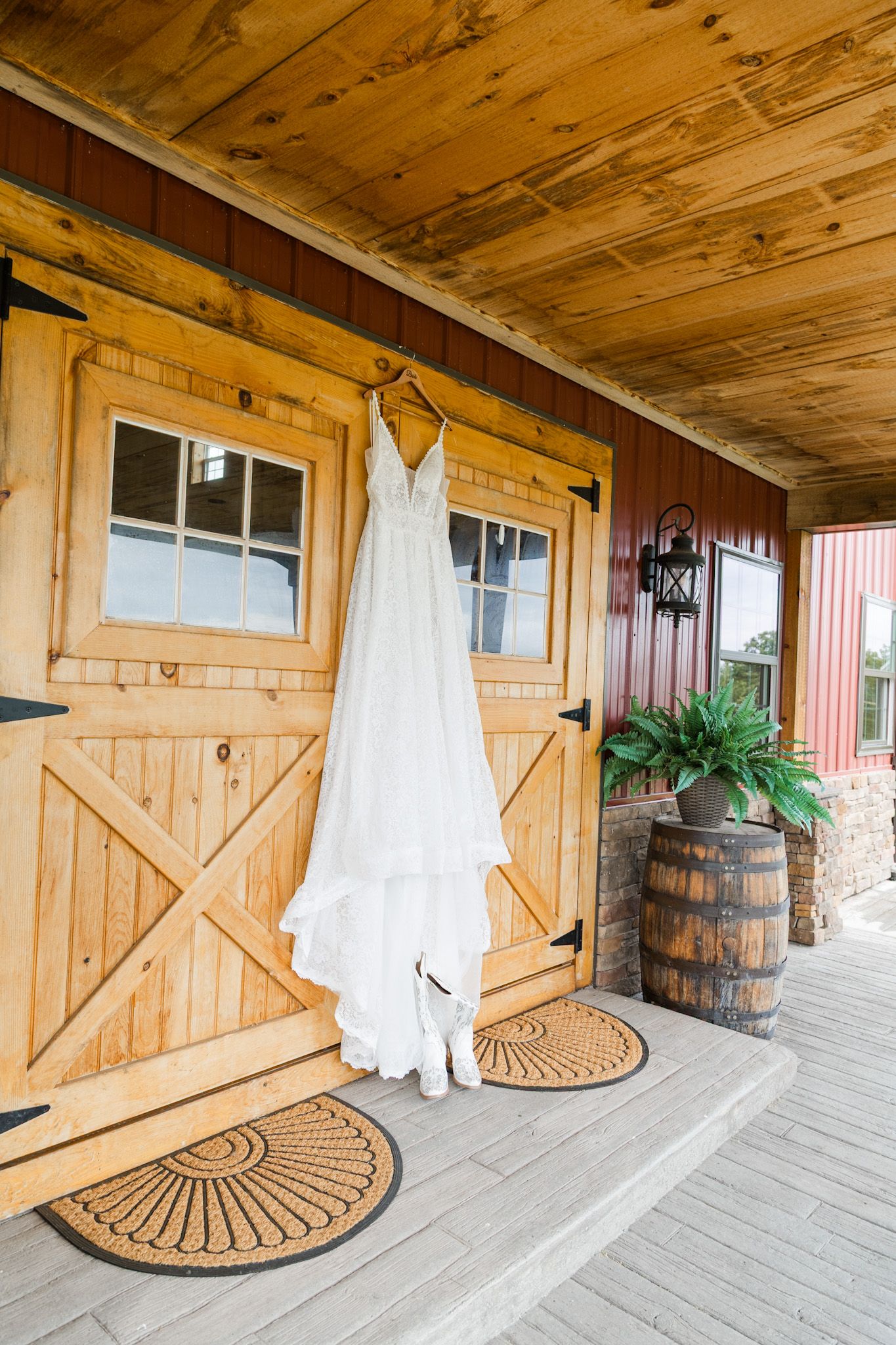White wedding dress hanging on rustic barn doors with cowboy boots below on a wooden porch with barrel decor.