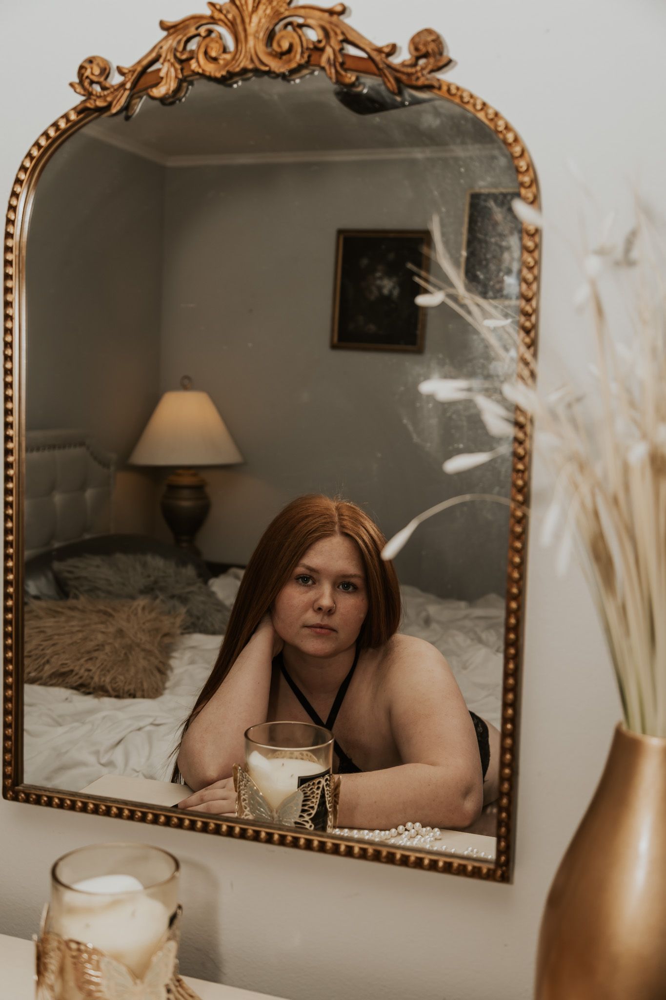 Redhead woman in black lingerie reflected in ornate gold mirror on dresser.