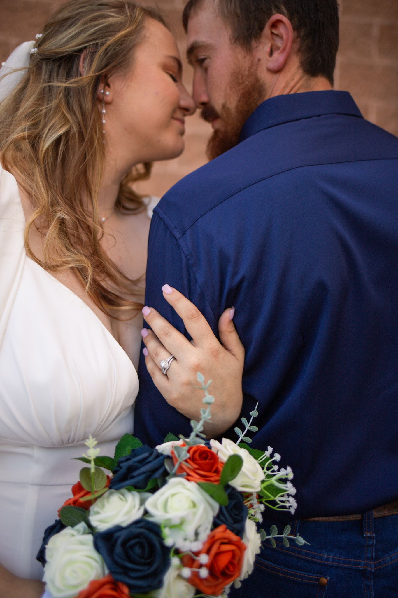 Bride and groom sharing romantic almost-kiss, bride holding navy and orange rose bouquet against brick wall.