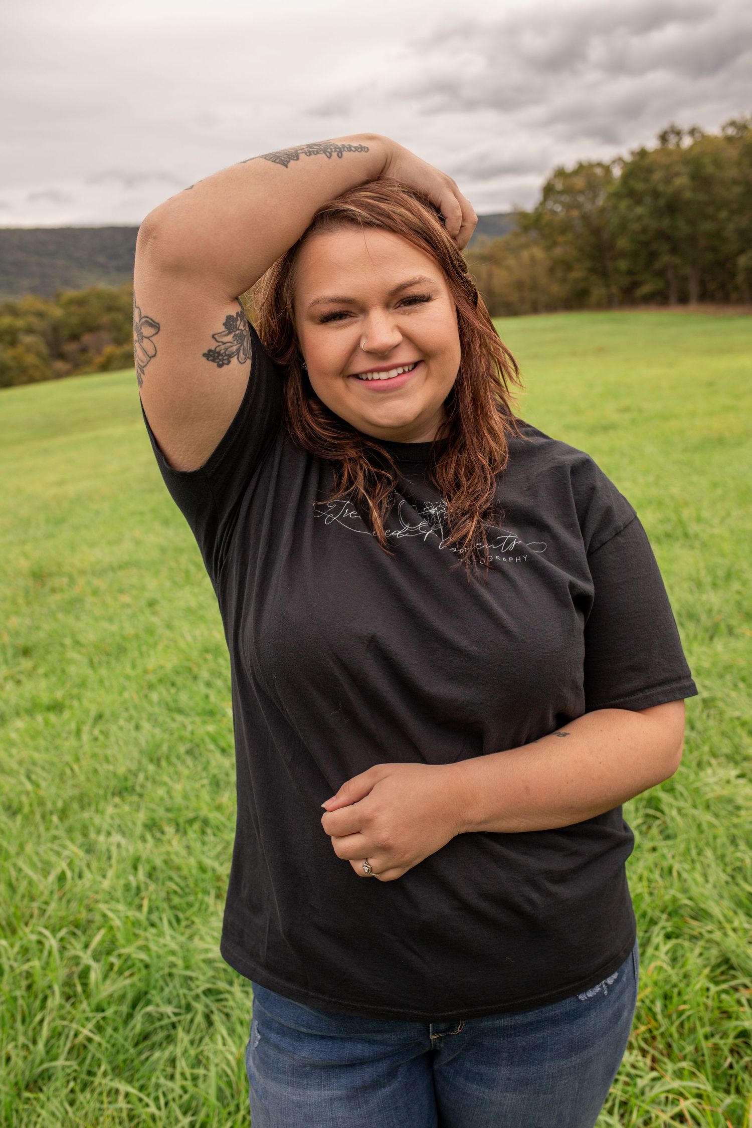 Tattooed woman in black tee smiling in green meadow under cloudy sky.