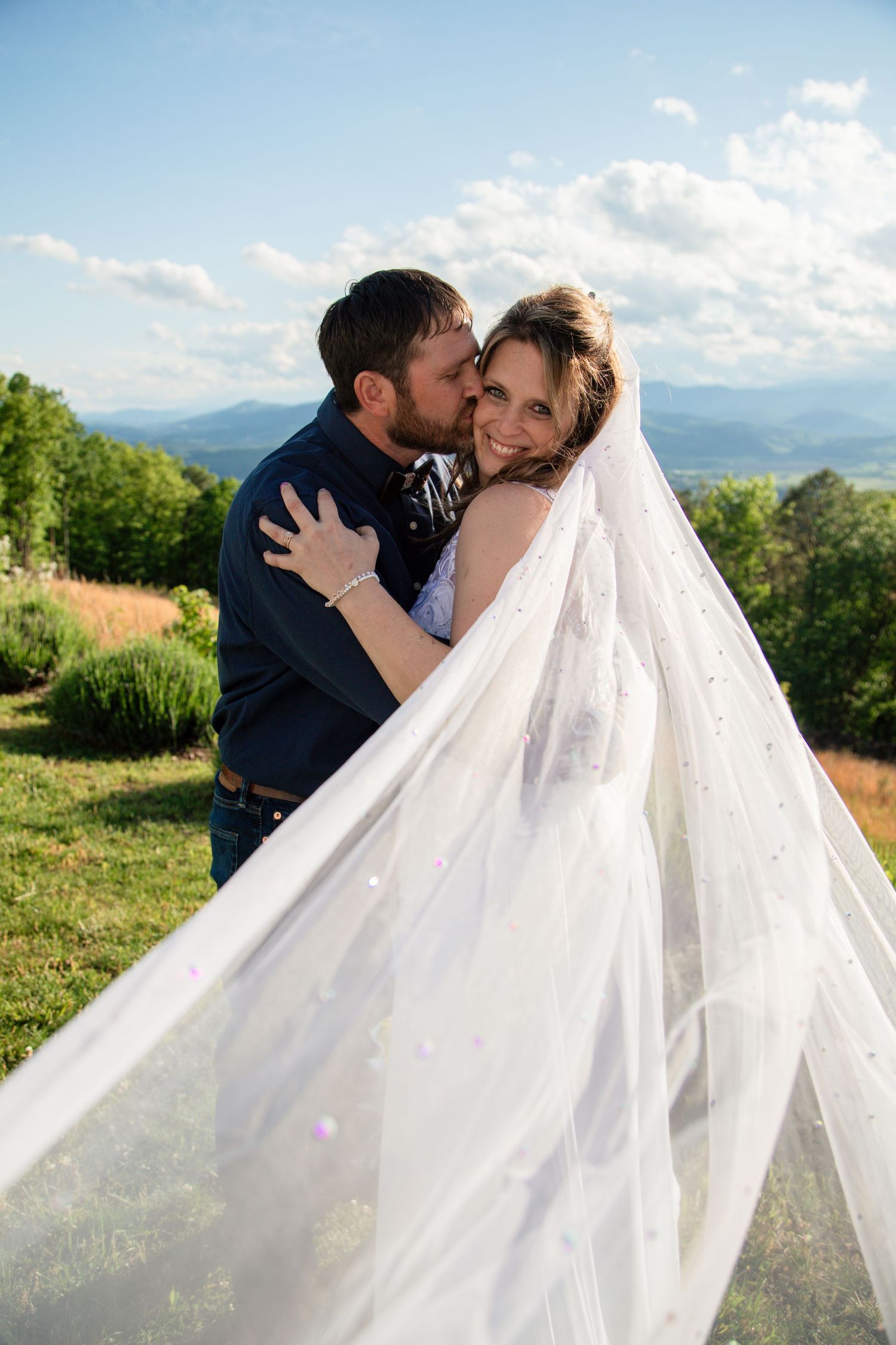 Groom kissing bride on cheek with flowing veil and stunning mountain landscape in background.