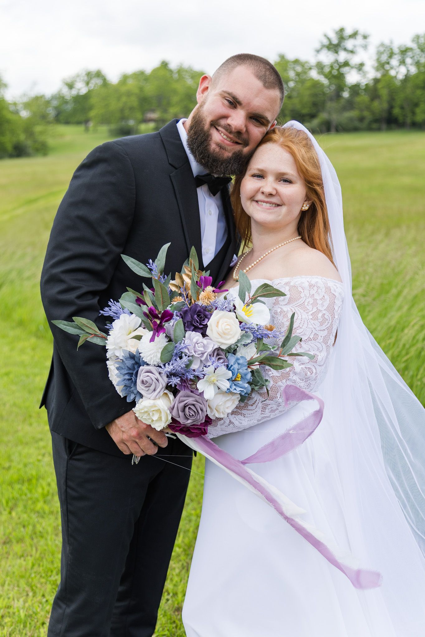 Happy couple posing in green field, groom in black tuxedo, bride holding purple and white bouquet.
