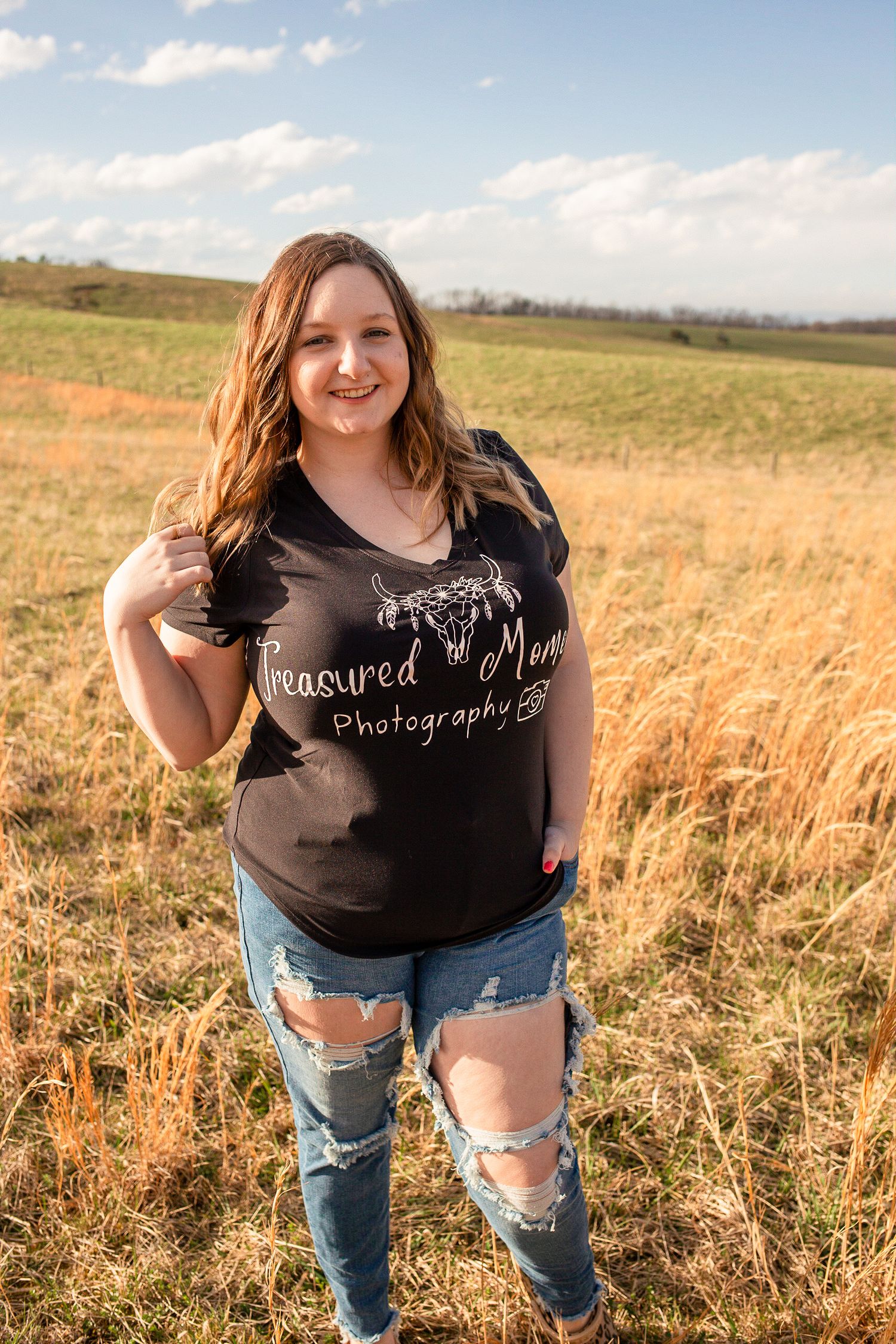 Smiling brunette woman in a black Treasured Moments Photography shirt and ripped jeans posing in a sunny field.