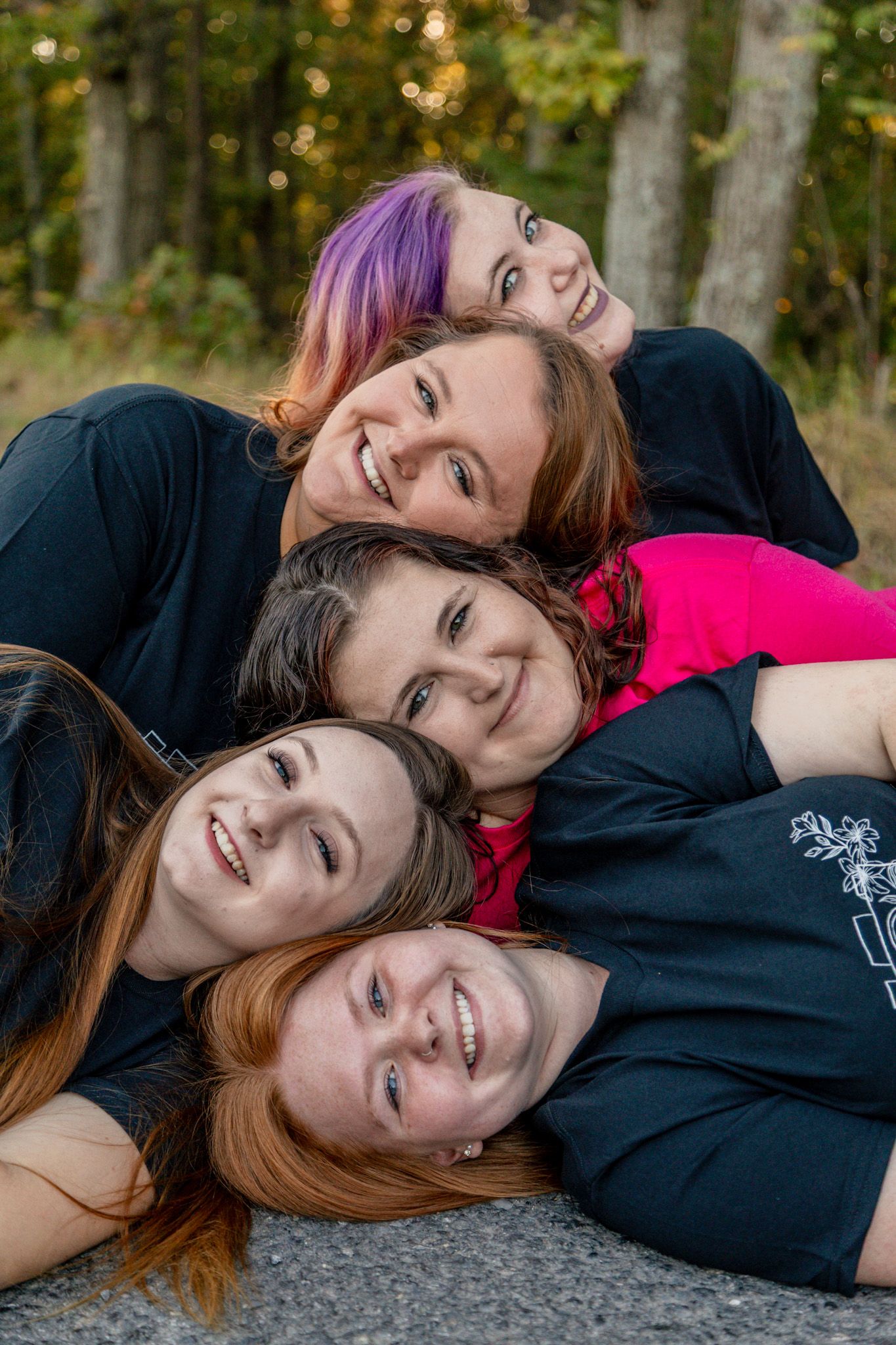 Five smiling women stacked together lying on the ground with colorful autumn trees behind them.