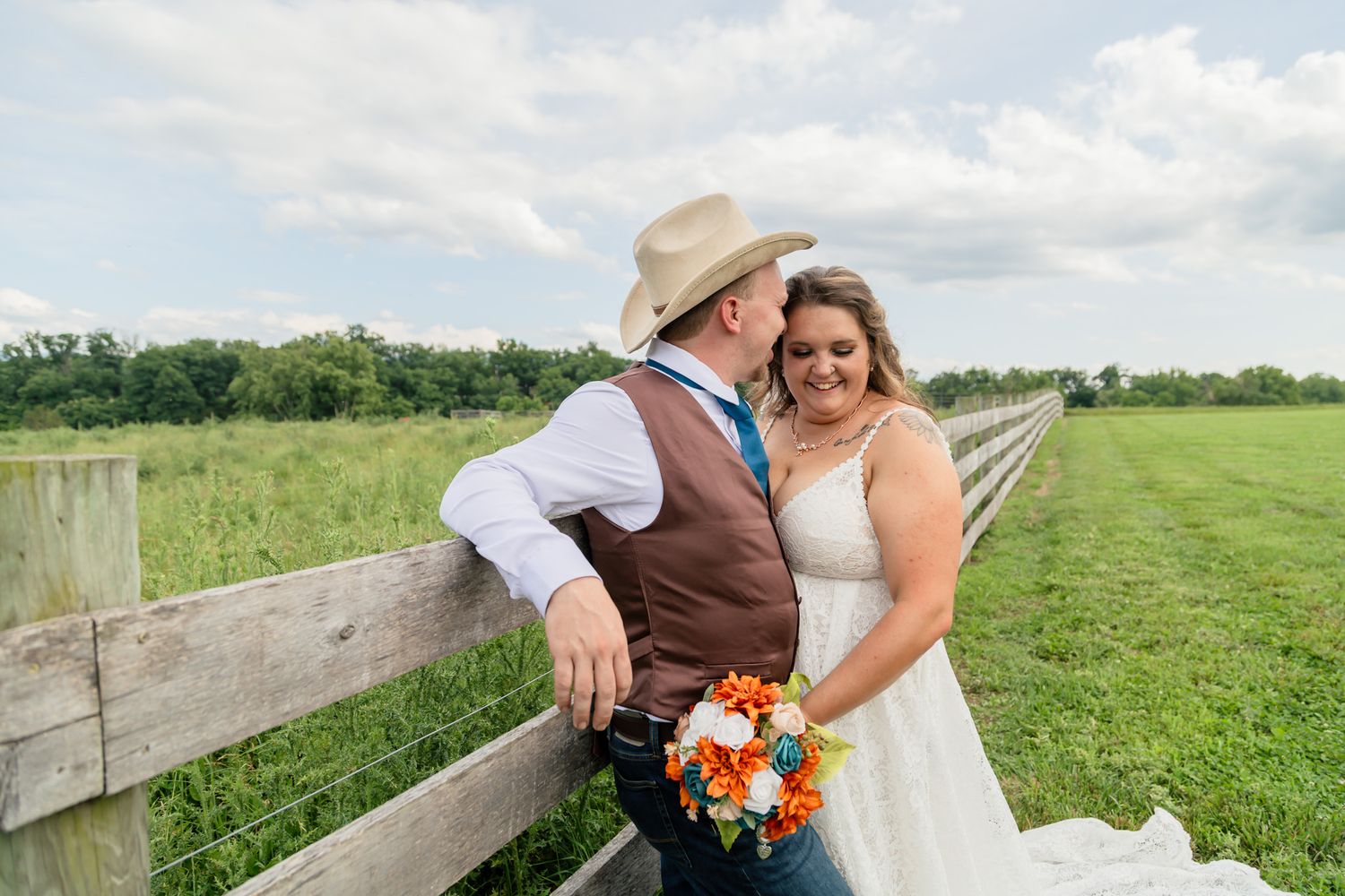 Bride and groom share a romantic moment by a rustic wooden fence in a green countryside field.
