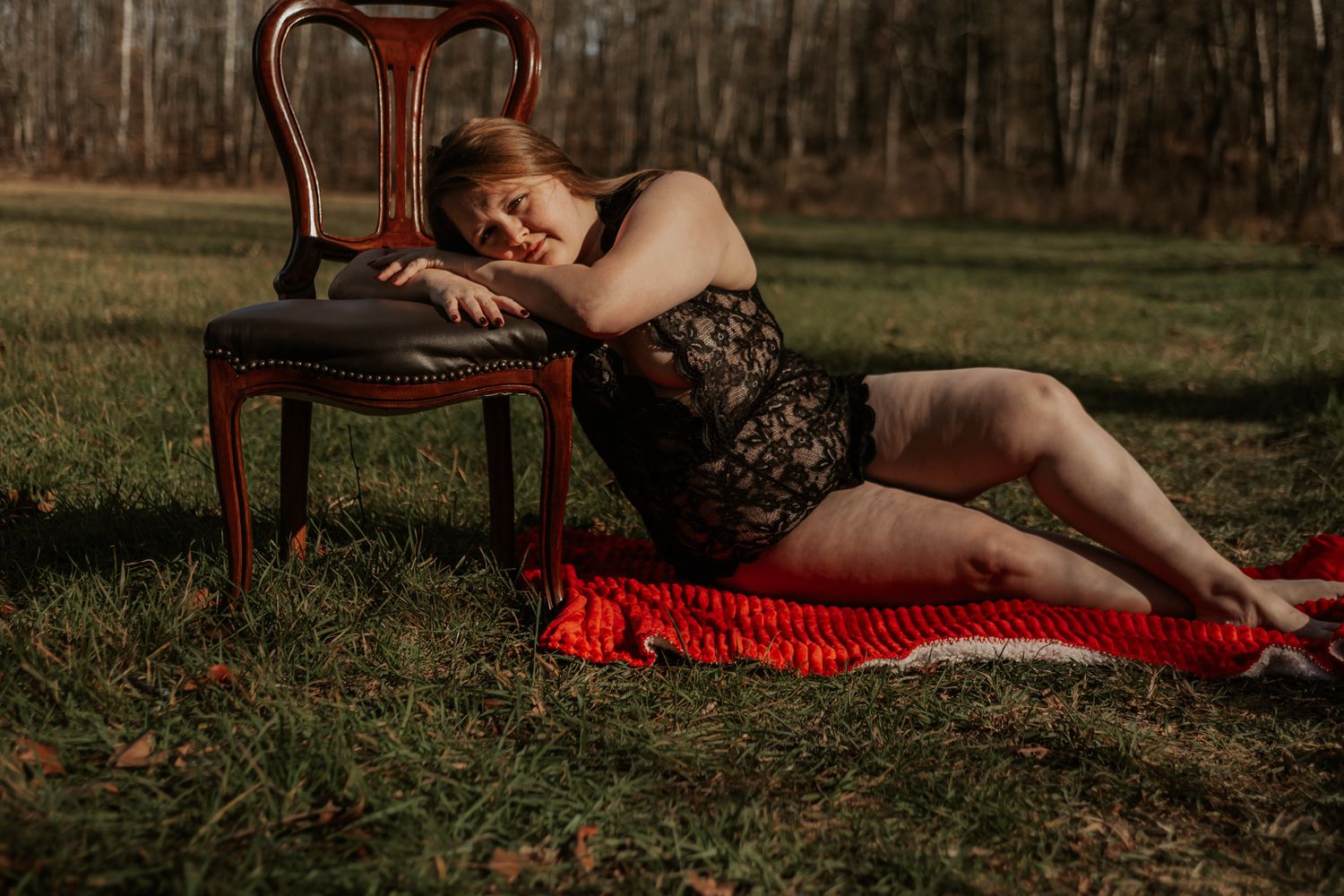 Woman in black lace bodysuit reclines on red blanket beside antique chair in autumn field.