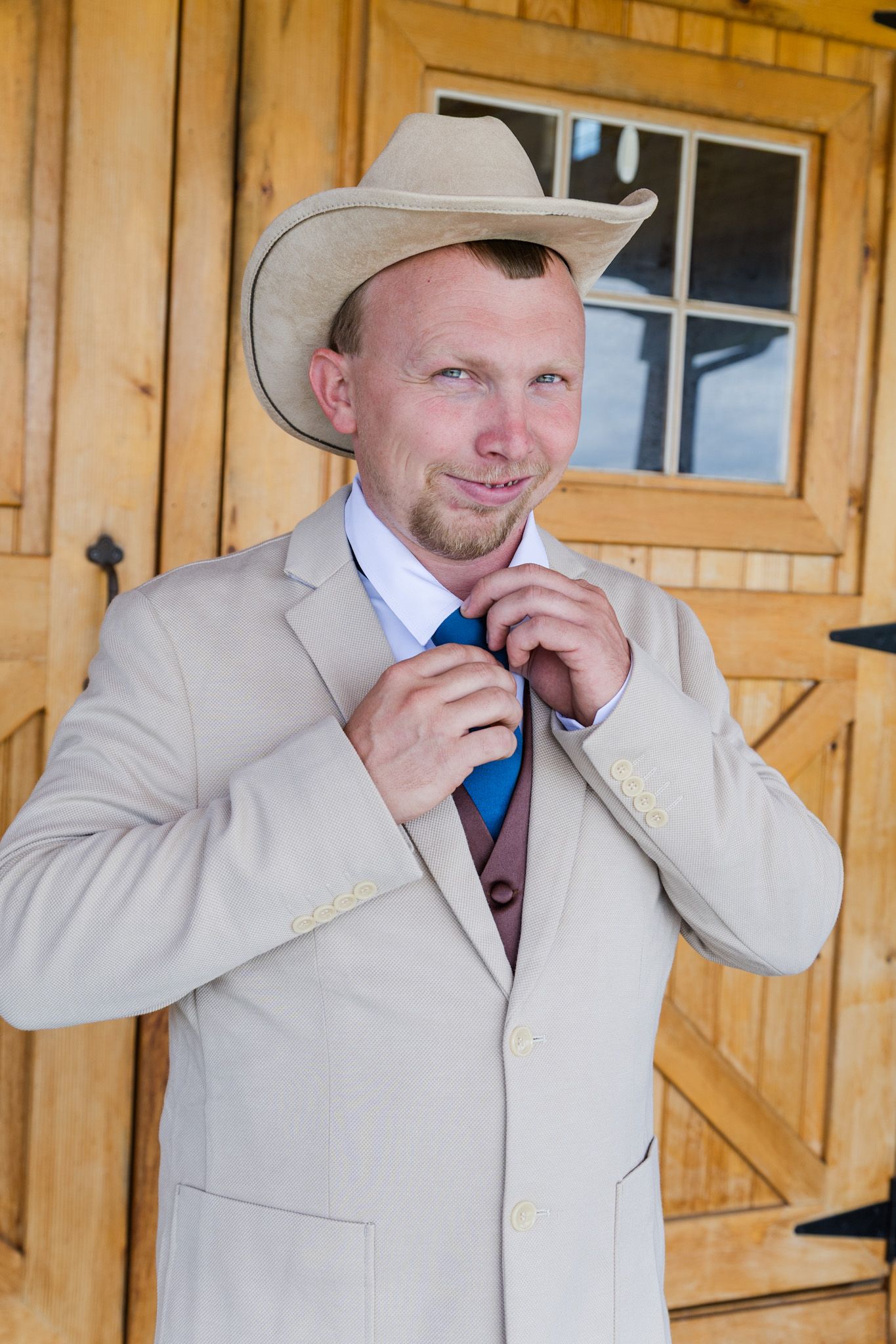 Groom in tan suit and cowboy hat adjusts blue tie outside rustic wooden barn doors.