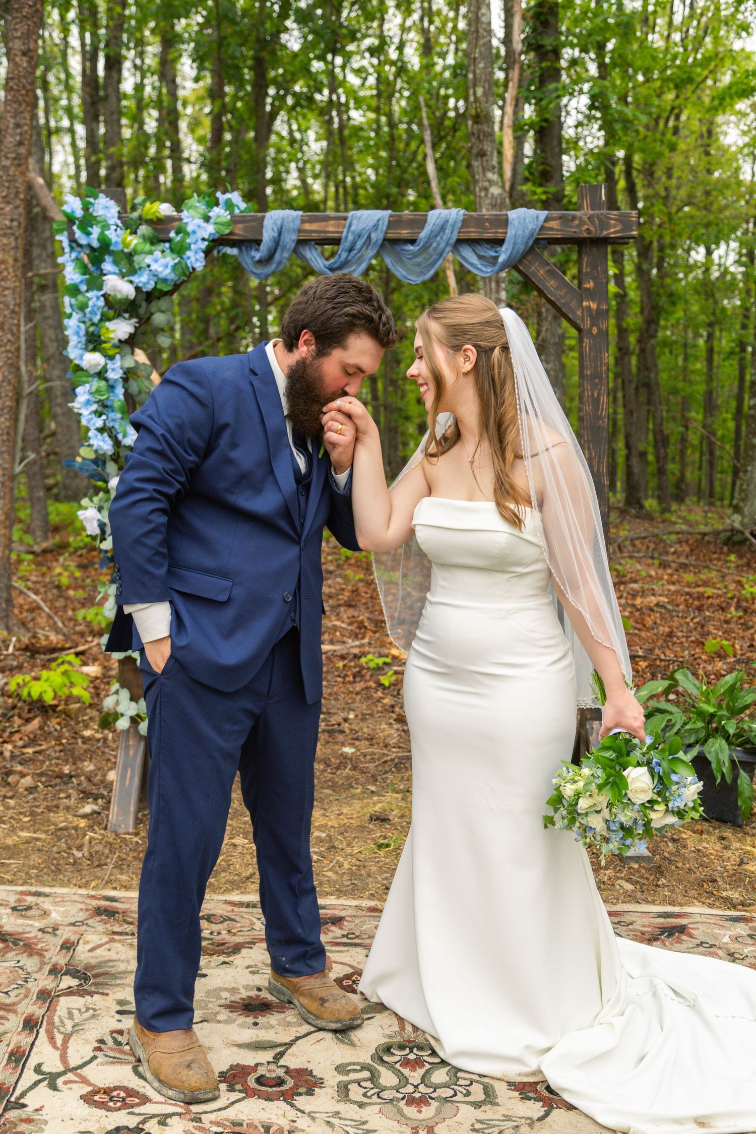 Groom in navy suit kisses bride's hand at outdoor woodland wedding ceremony with blue floral arch.