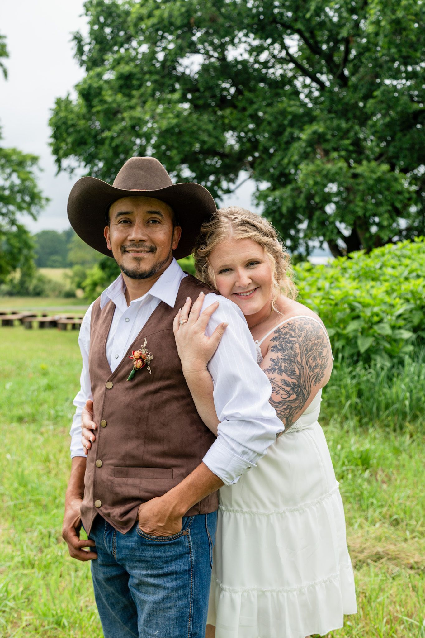 Smiling couple posing outdoors, groom in cowboy hat and brown vest, bride in white dress with tattoos.