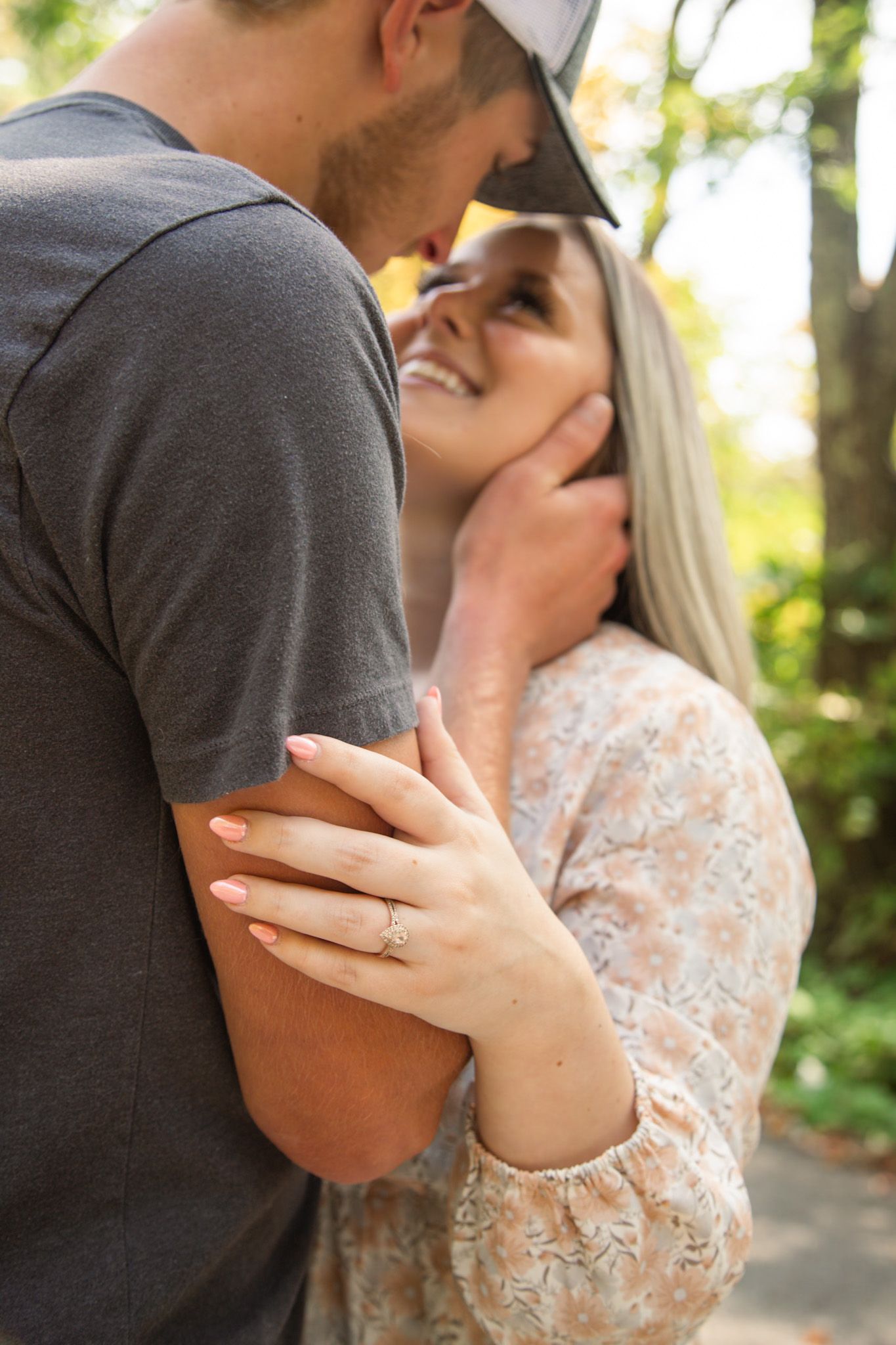 Smiling woman with engagement ring embracing man tenderly in a sunlit outdoor park setting.