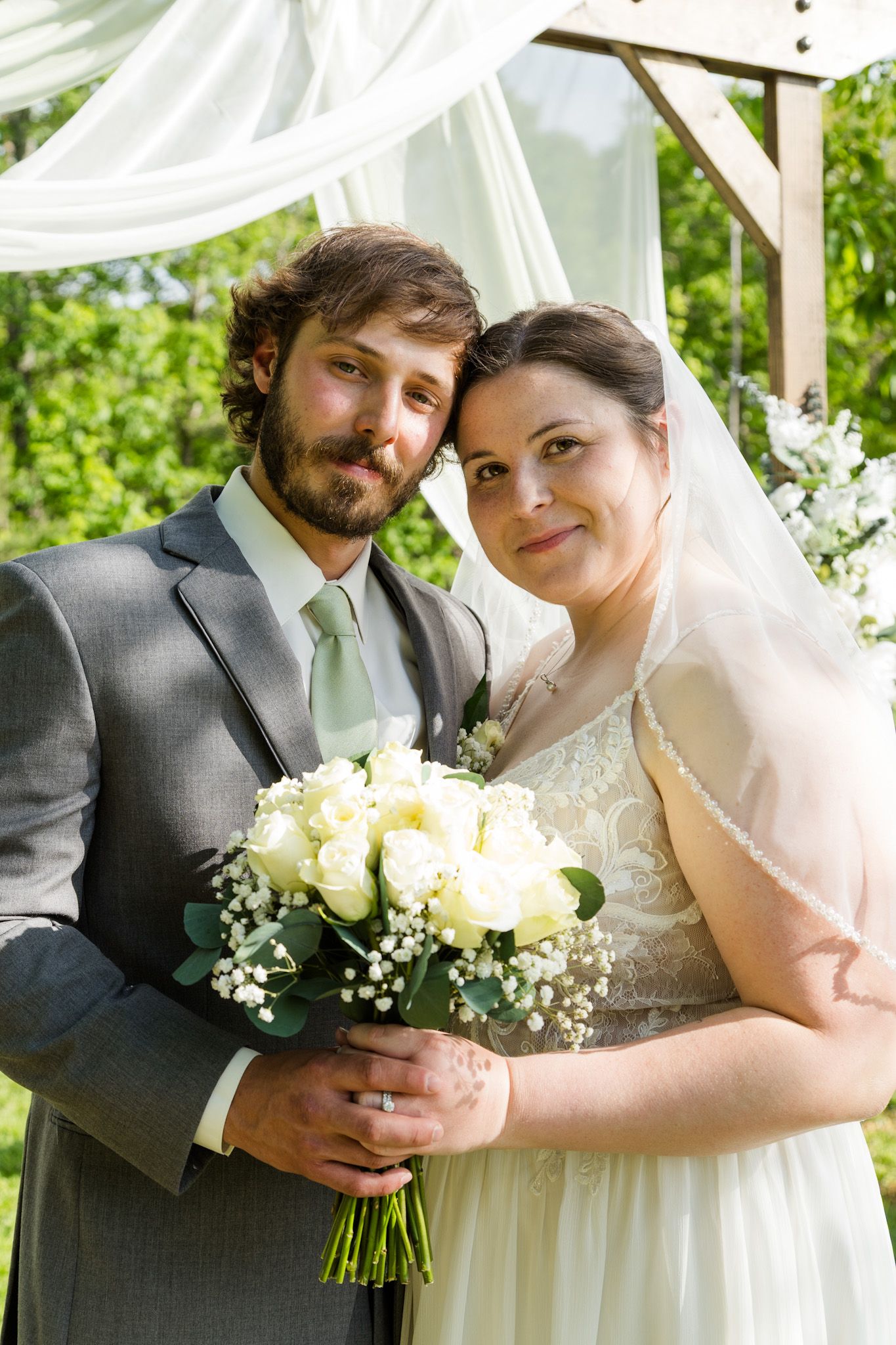 Bride and groom pose together outdoors holding white rose bouquet under decorated wooden arch.