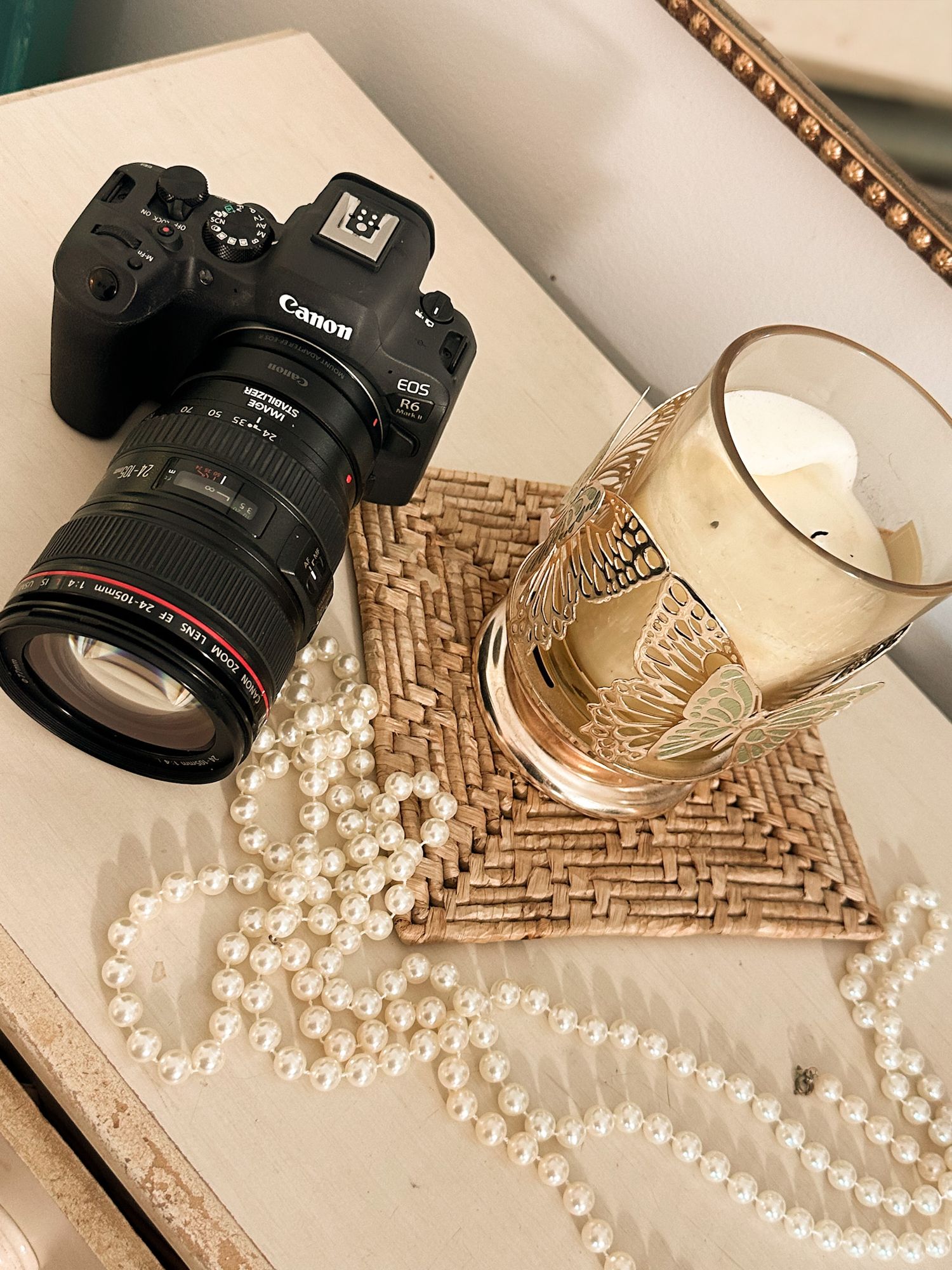 Canon EOS camera with lens beside pearl necklace and gold candle on decorative tray.