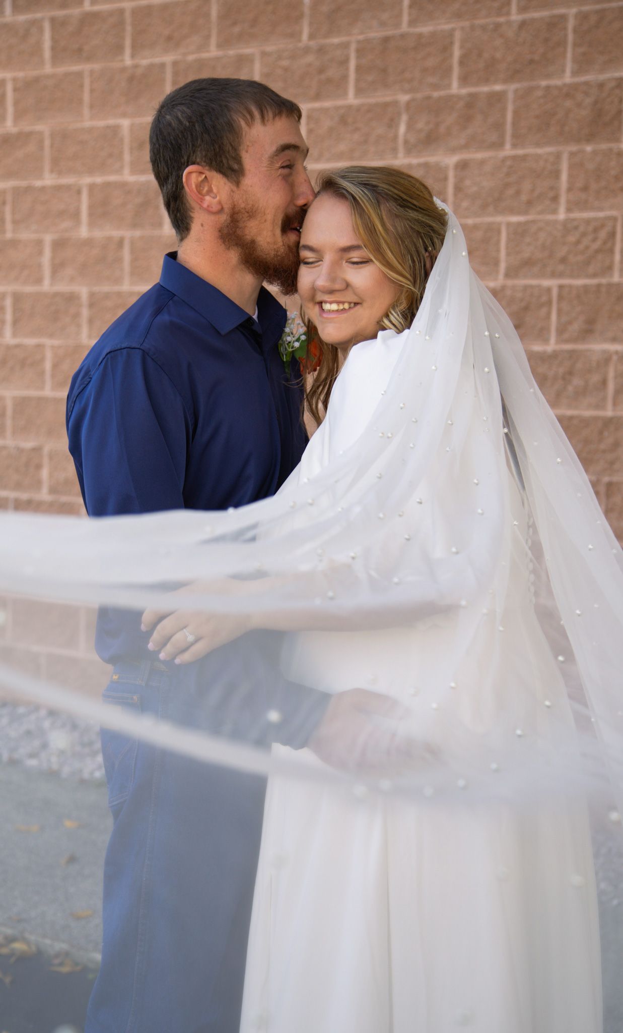 Smiling bride in white veil embraced by groom in blue shirt against brick wall.