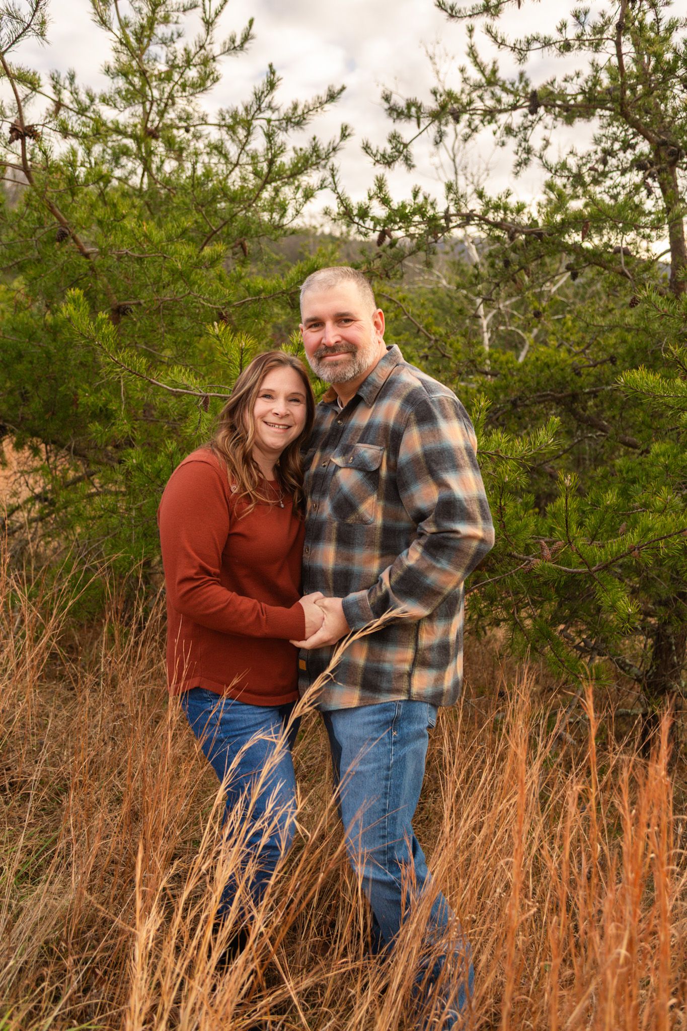 Couple holding hands and smiling in autumn field with pine trees and golden grass in Colorado mountains.