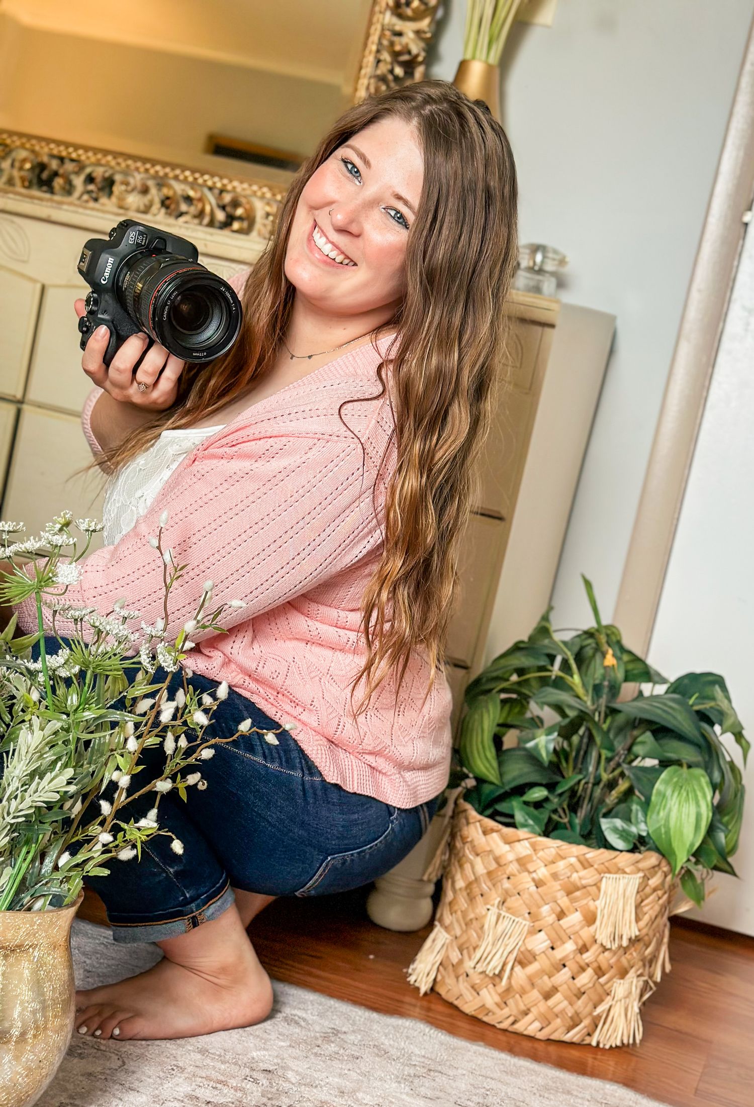 Smiling woman crouching indoors holding a camera, surrounded by potted plants in a bright decorated room.