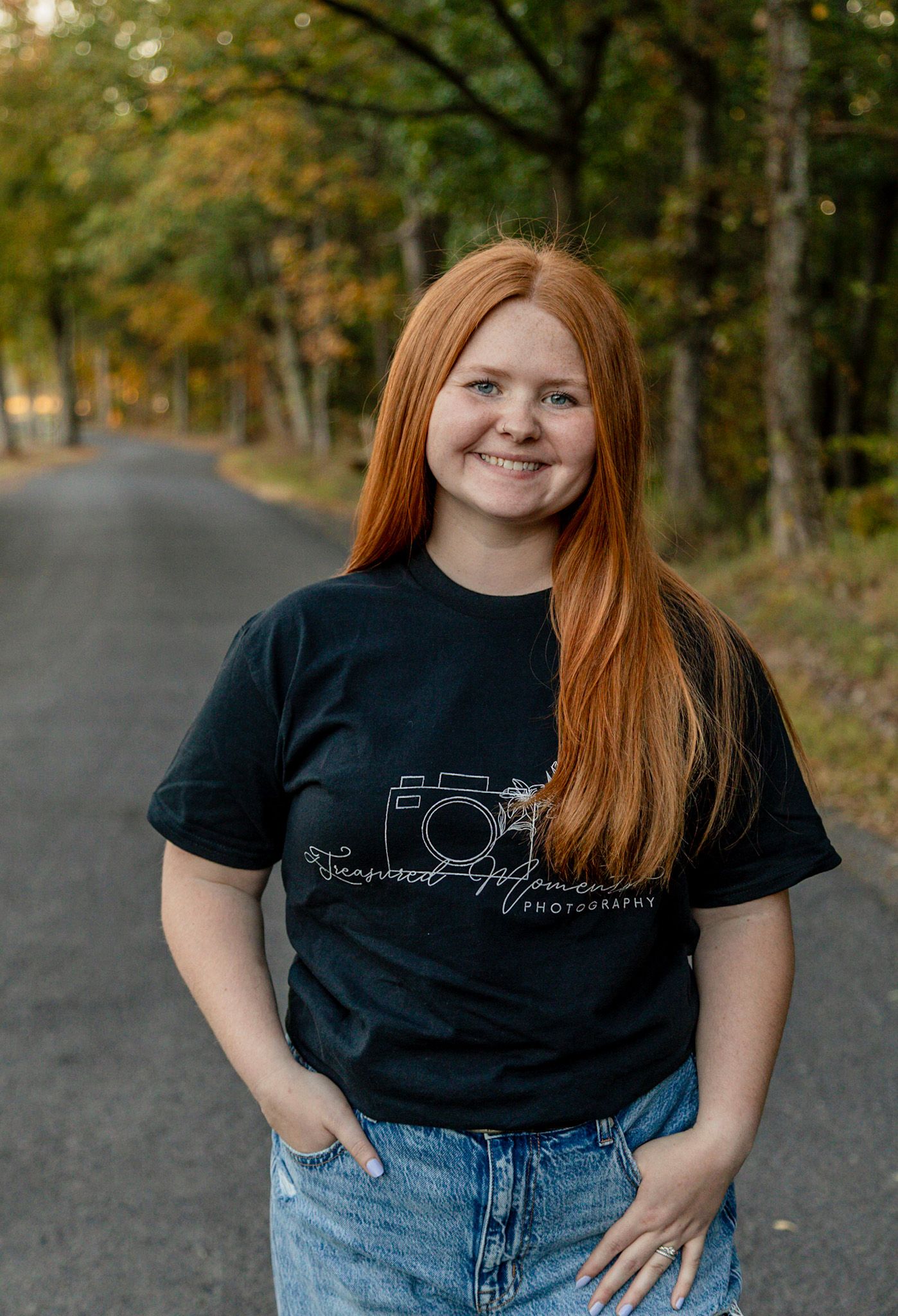 Young woman with long red hair smiling outdoors, wearing a black photography branded t-shirt on a tree-lined road.