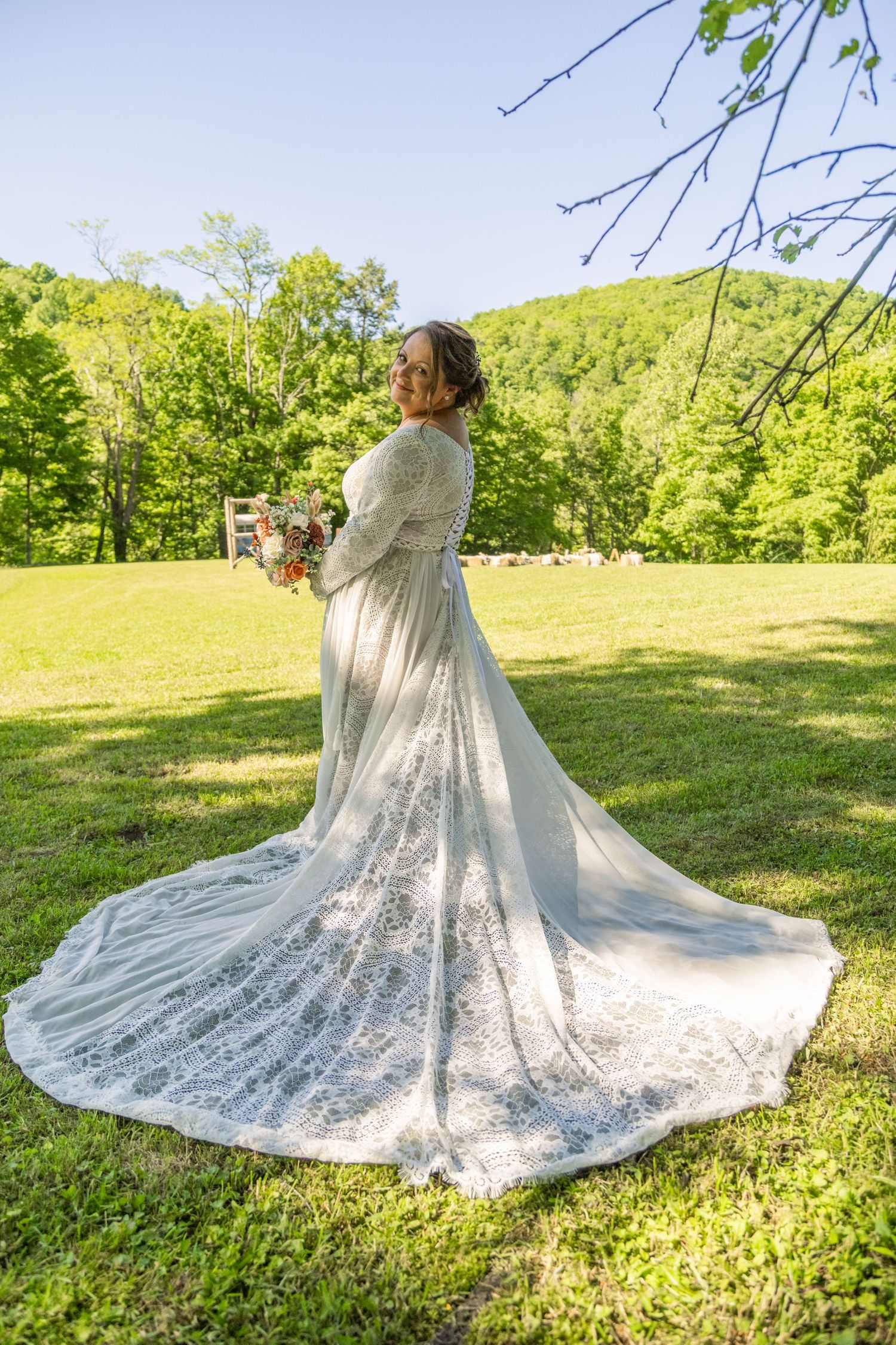 Bride in elegant long-sleeve lace wedding gown with train stands in green meadow holding bouquet, forest hills backdrop.