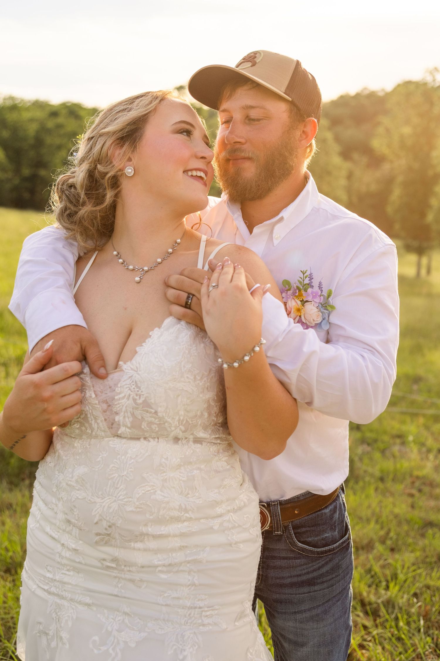 Bride and groom sharing a romantic moment outdoors at golden hour, she in lace gown, he in white shirt and cap.