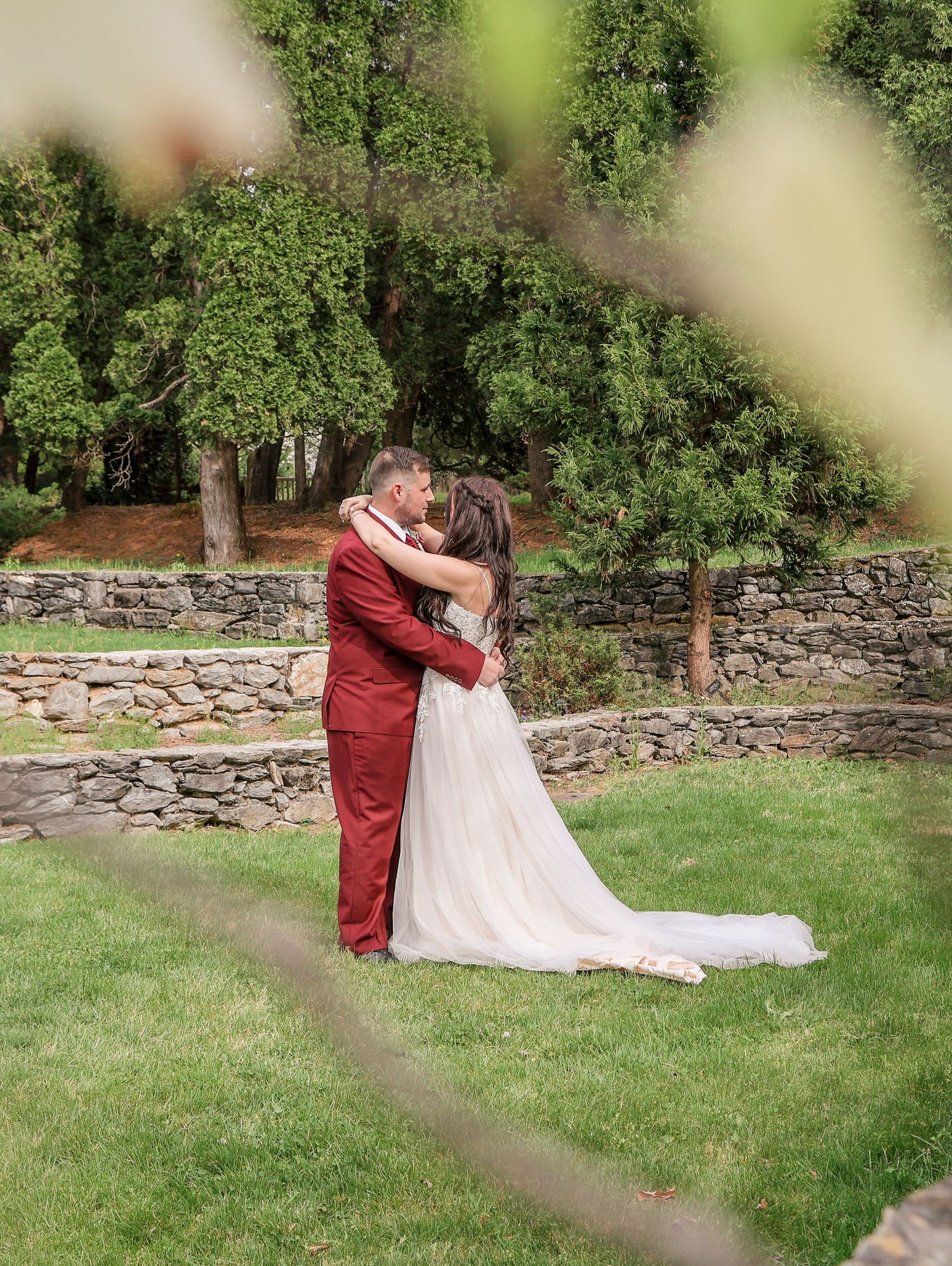 Bride and groom embrace and kiss on lush green lawn with stone wall backdrop.