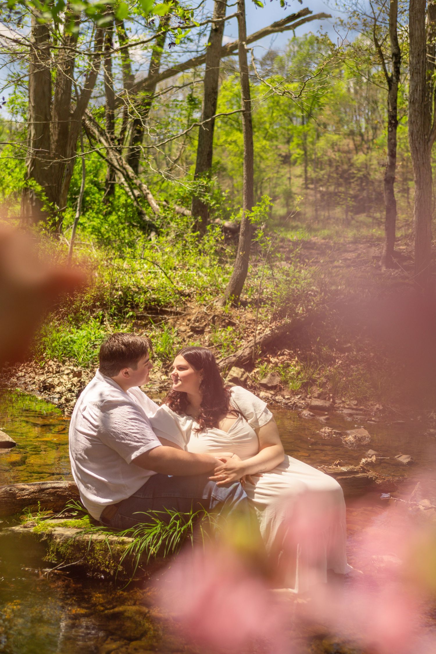 Pregnant couple sitting by a sunlit forest creek, sharing a tender moment surrounded by lush green trees and spring blooms.