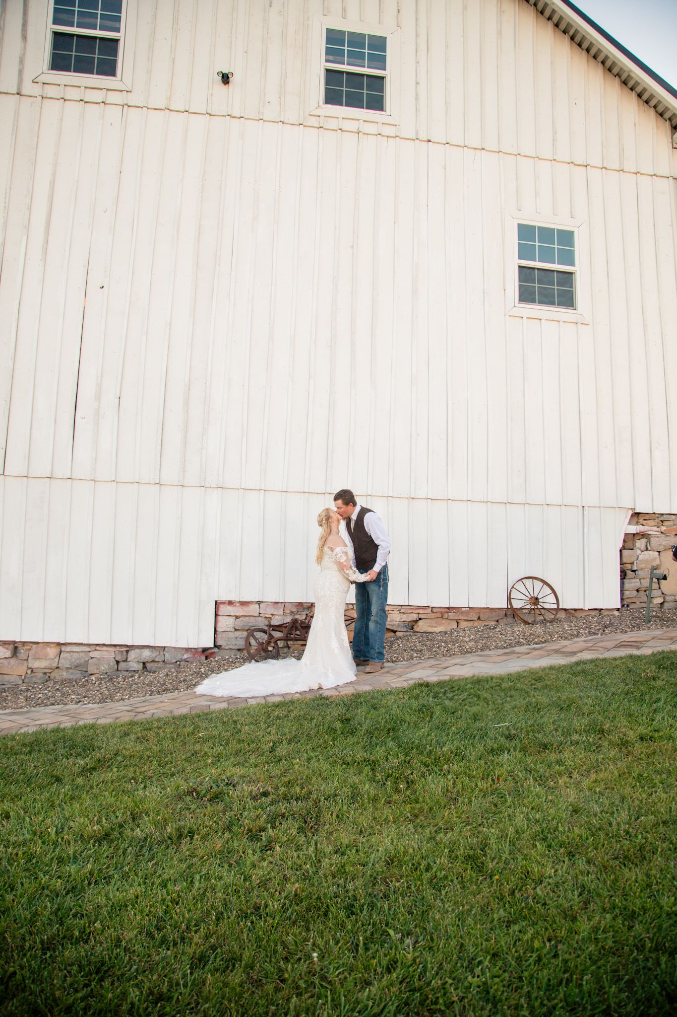 Bride and groom kissing beside large white barn with stone foundation on wedding day.