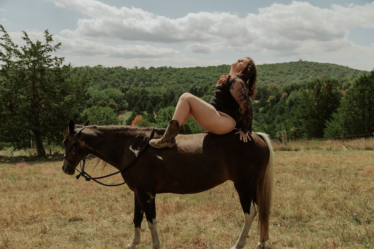 Tattooed woman in black bodysuit posing on horseback in a scenic open field with forest backdrop.