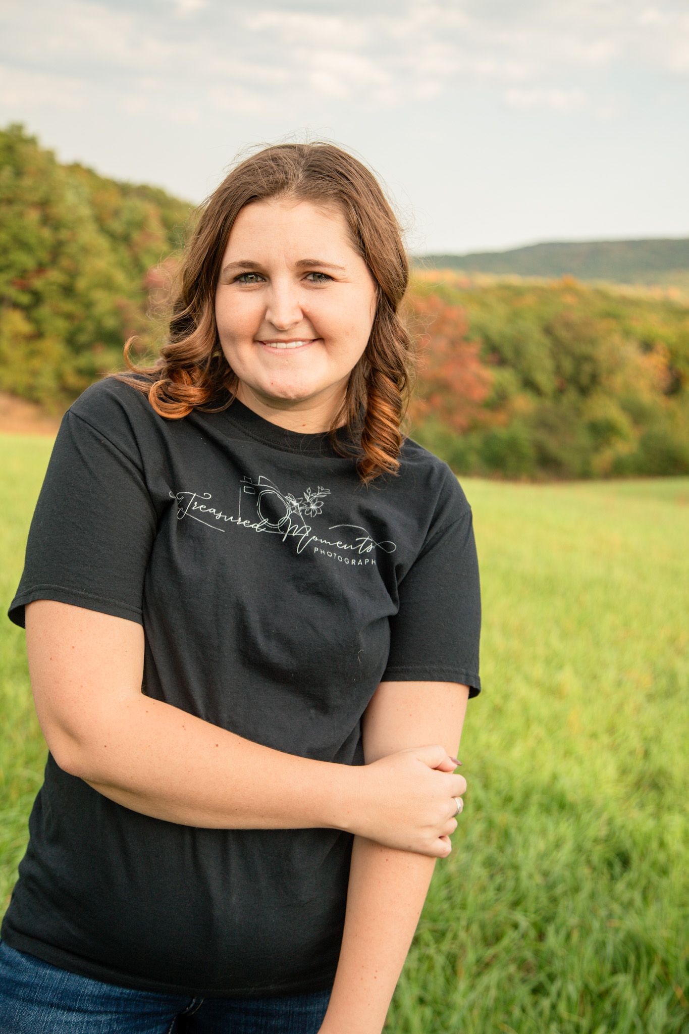 Young woman in black tee smiling with arms crossed in green field.
