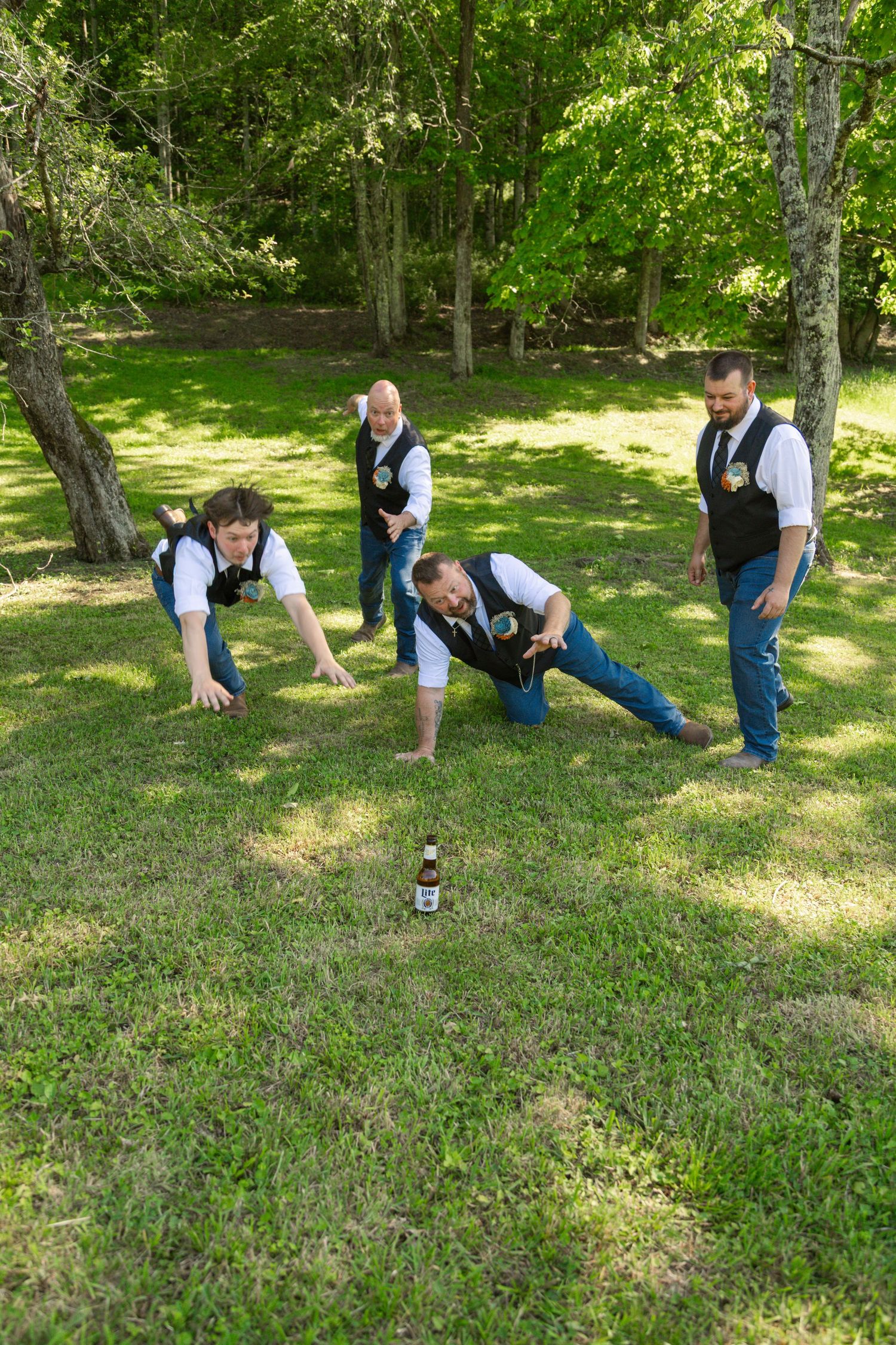 Four groomsmen in vests and jeans playing a lawn game with a beer bottle on green grass surrounded by trees.