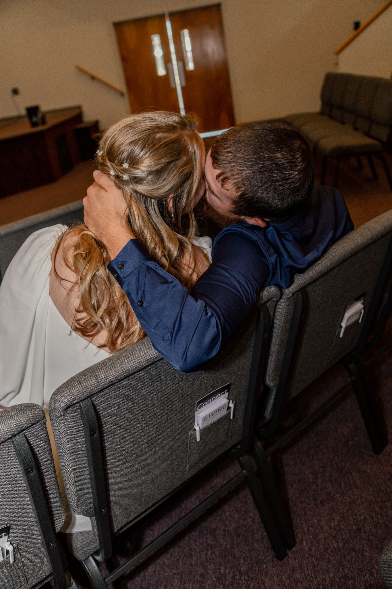 Bride and groom share intimate moment seated together in church pews from behind.