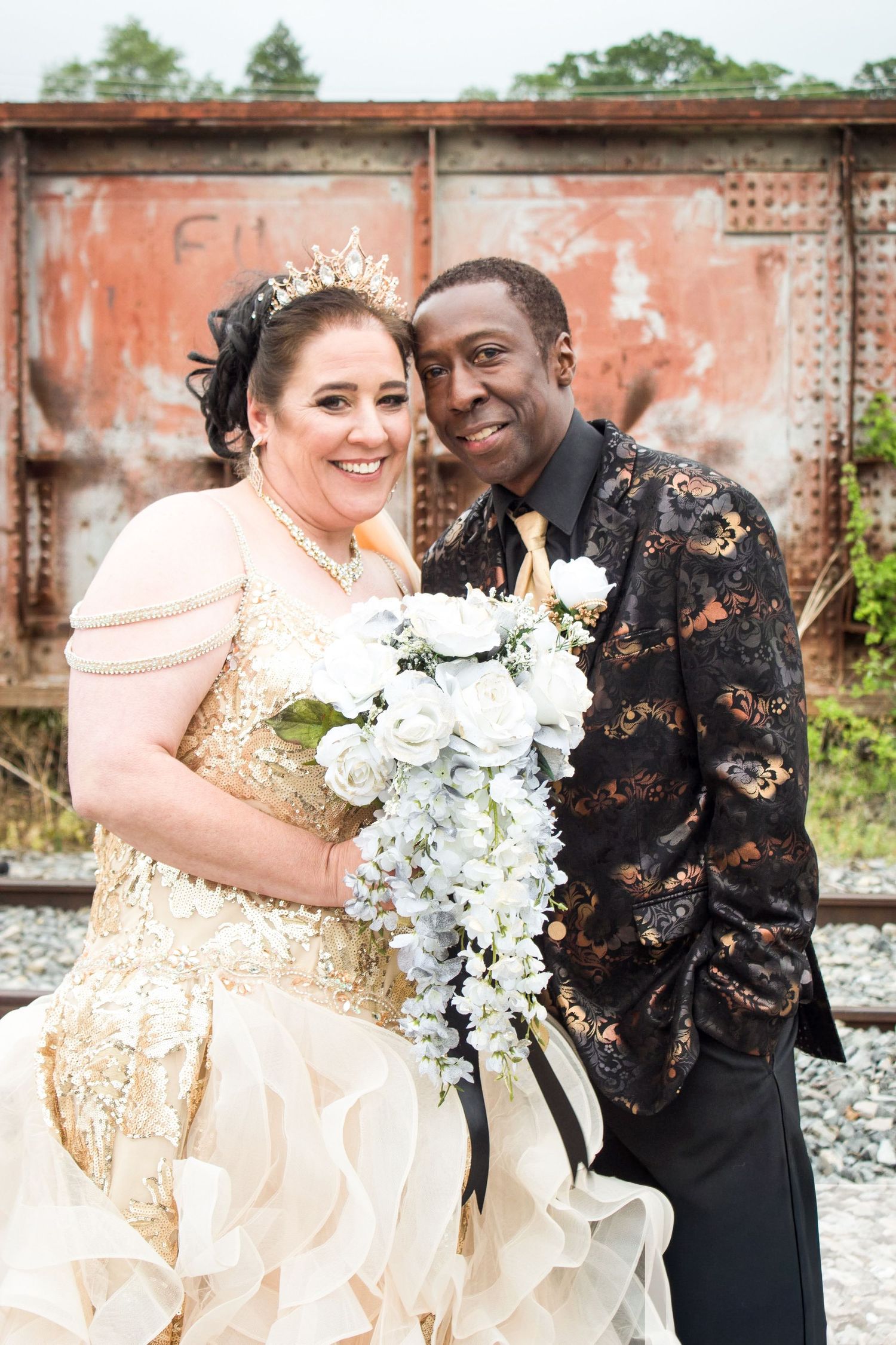 Interracial couple in elegant wedding attire pose with white floral bouquet near rustic train car.