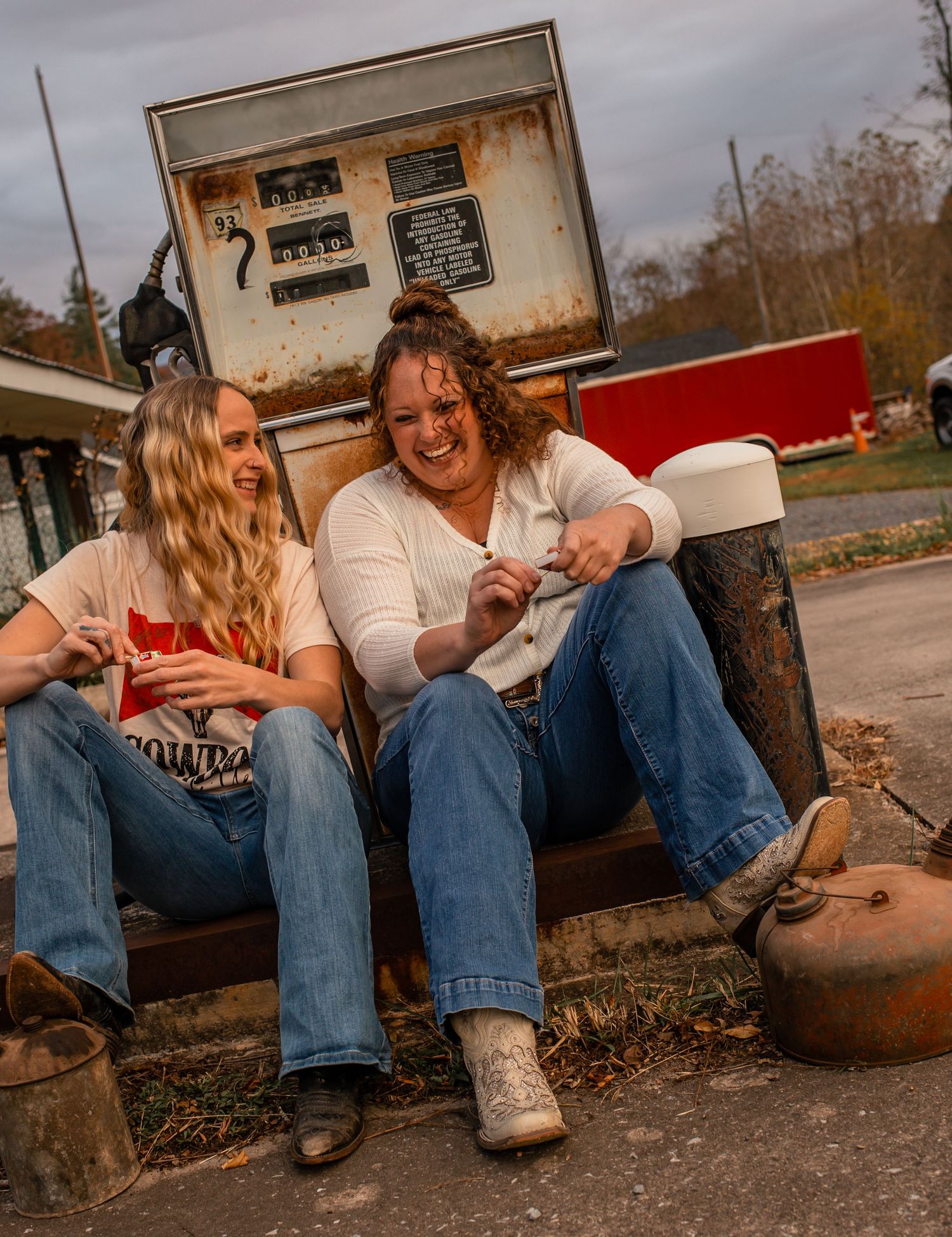 Two women laughing together while sitting near a rustic vintage gas pump on a sunny autumn day.
