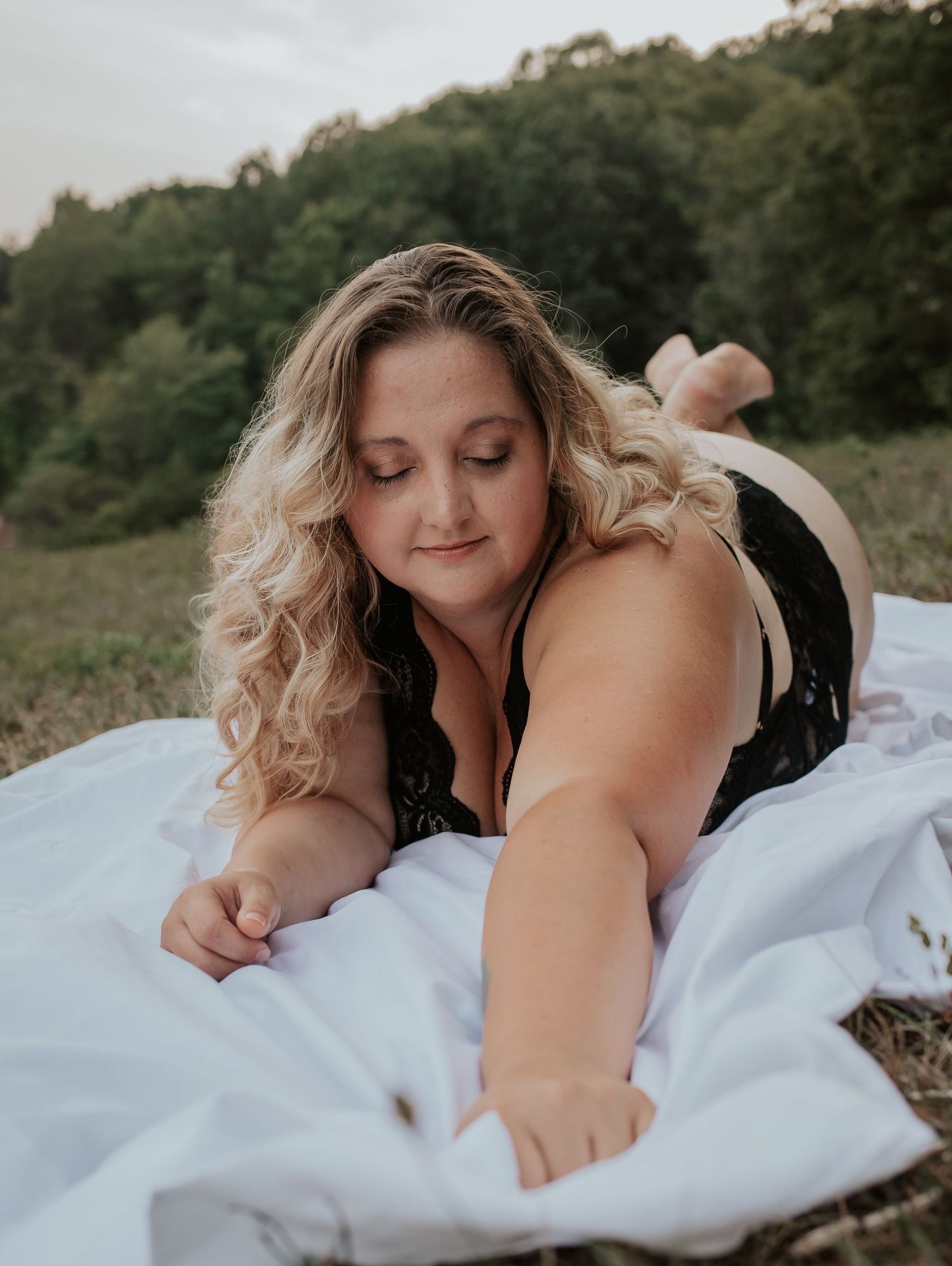 Curly blonde woman in black lace bodysuit lying on a white blanket in an open meadow.