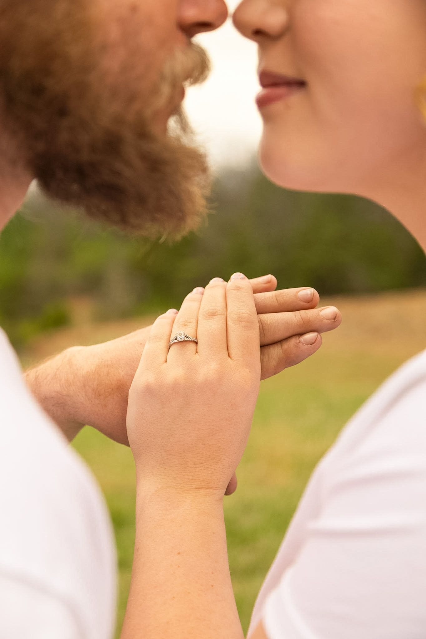 Close-up of a couple nearly kissing while holding hands, showcasing a diamond engagement ring outdoors.