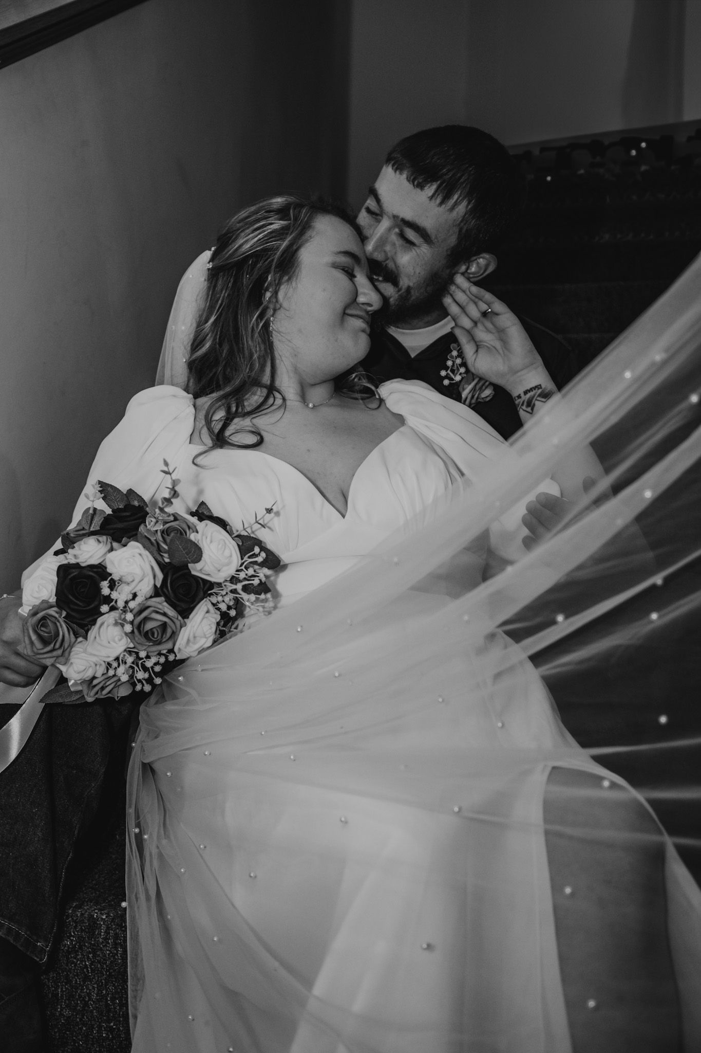 Black and white photo of bride with bouquet and groom sharing tender moment on stairs.