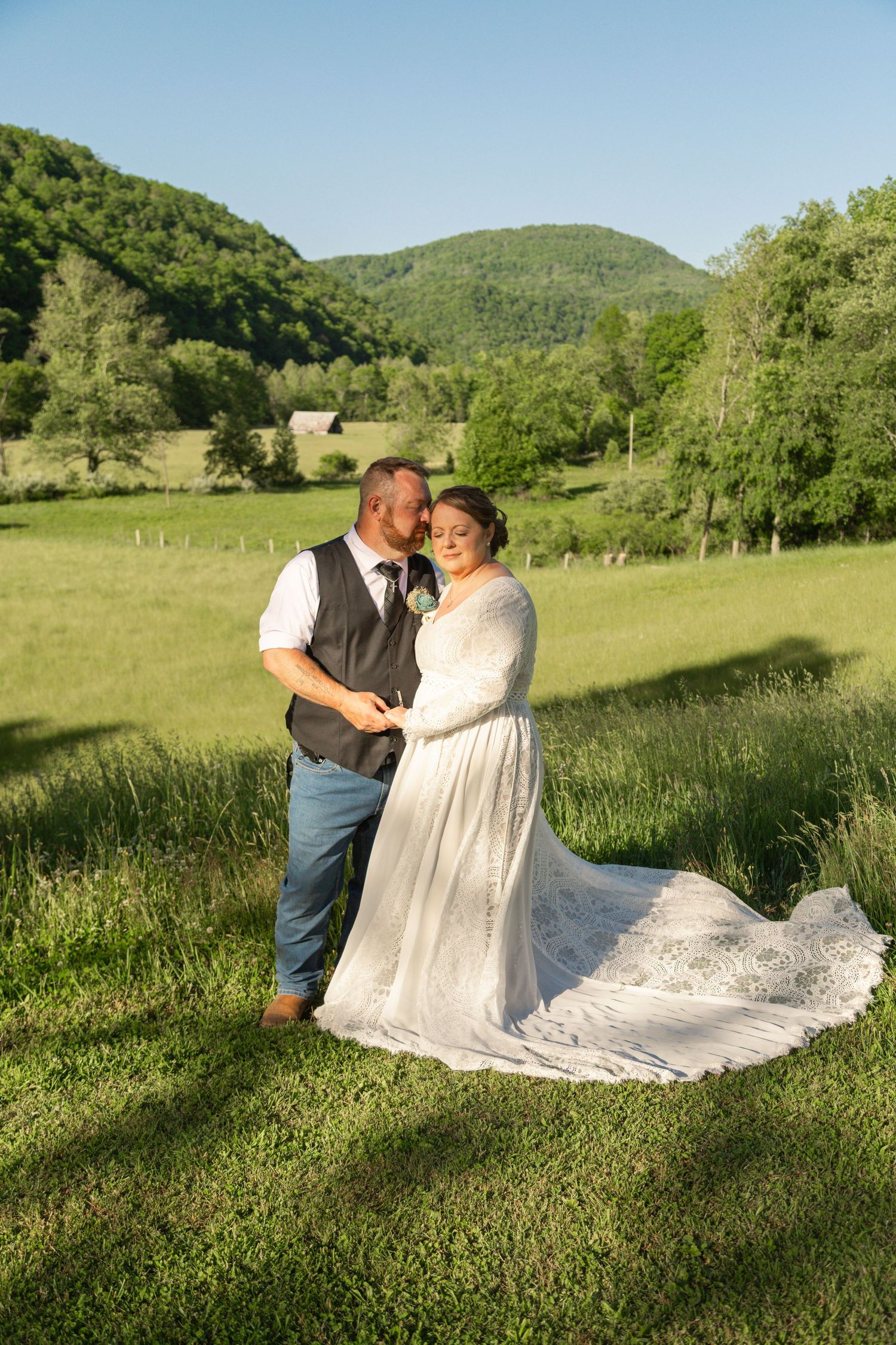 Bride in lace gown and groom in vest embrace in a sunlit green meadow with rolling mountains backdrop.
