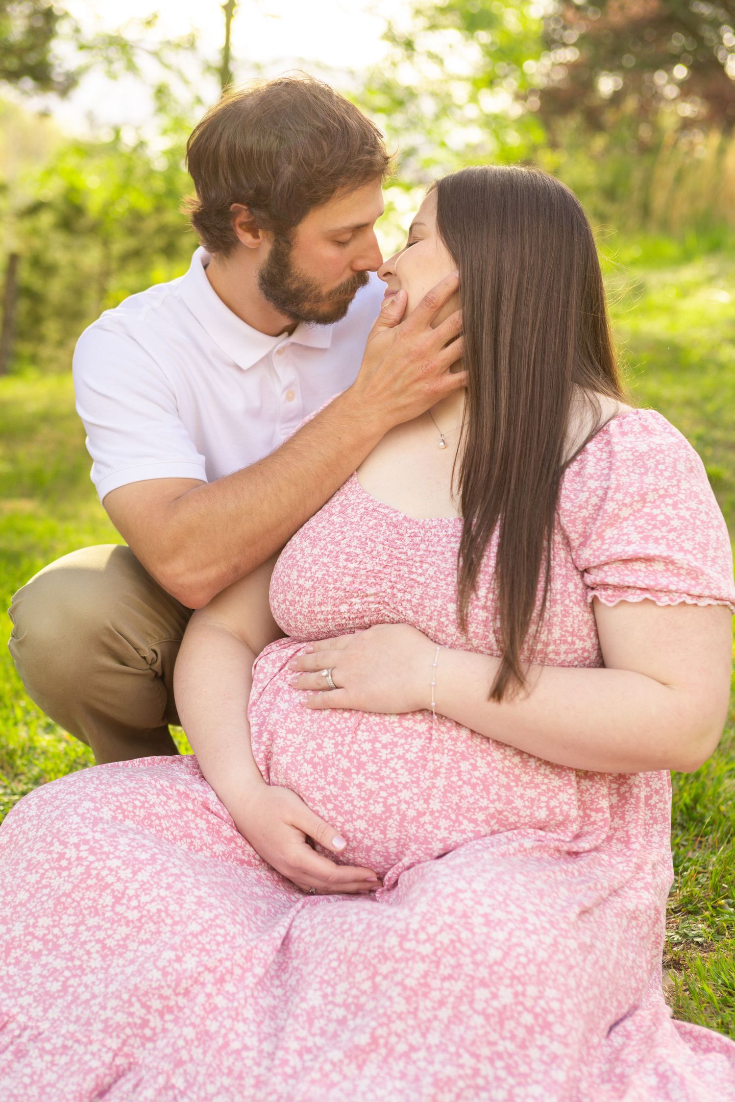 A couple shares a tender moment outdoors, with the pregnant woman in a pink floral dress cradling her baby bump.