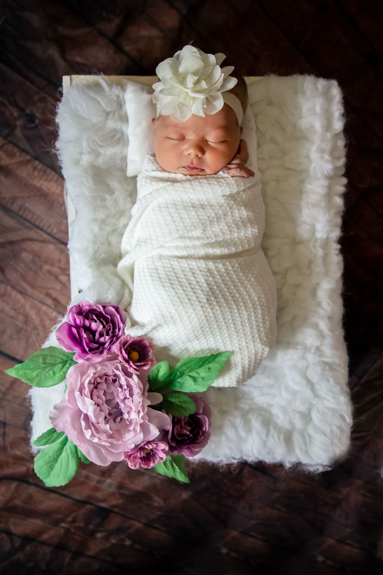 Sleeping newborn baby girl swaddled in white wrap with flower headband and purple floral decorations.