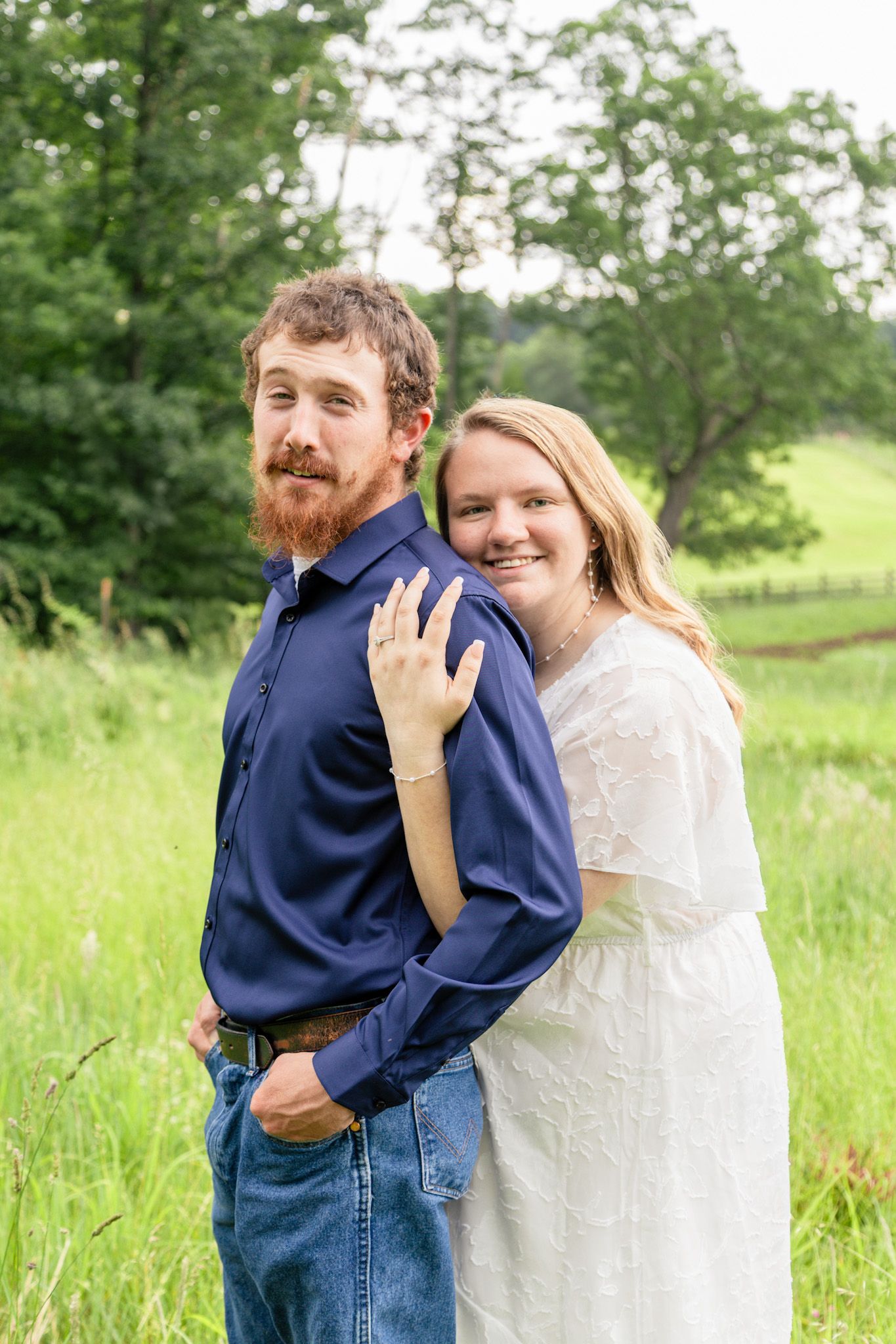 Smiling woman in white dress with fiance in blue shirt in green meadow.