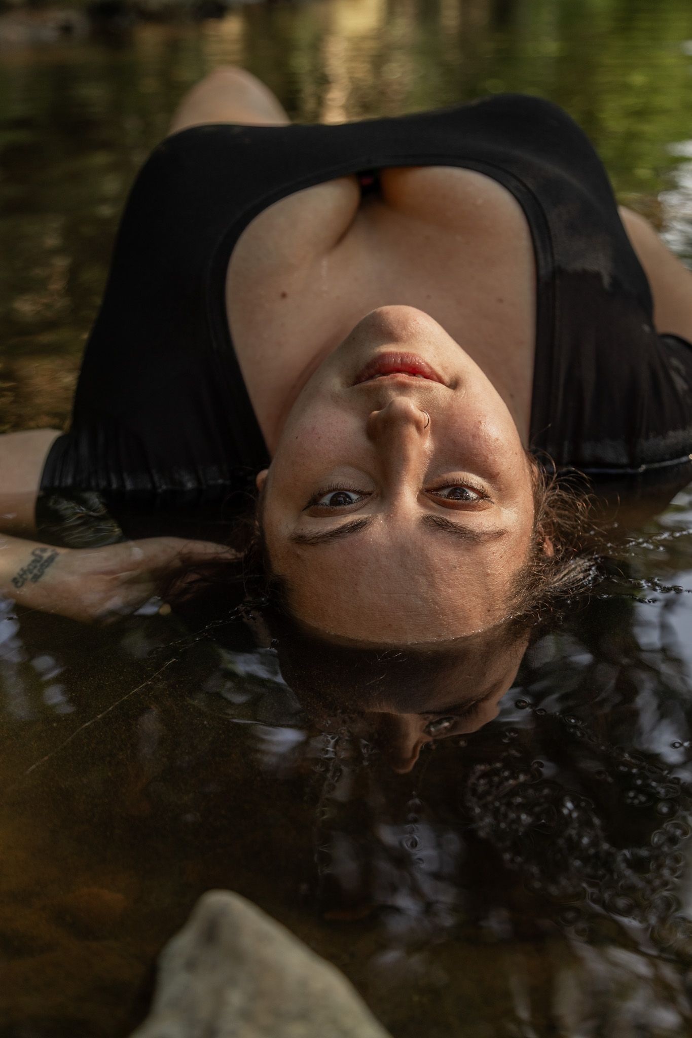 Woman in black dress lies in shallow creek water looking up at camera in forest setting.