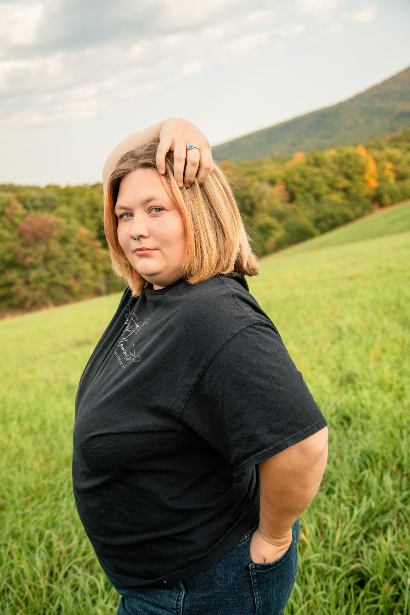 Blonde woman in black tee posing in scenic green autumn landscape.
